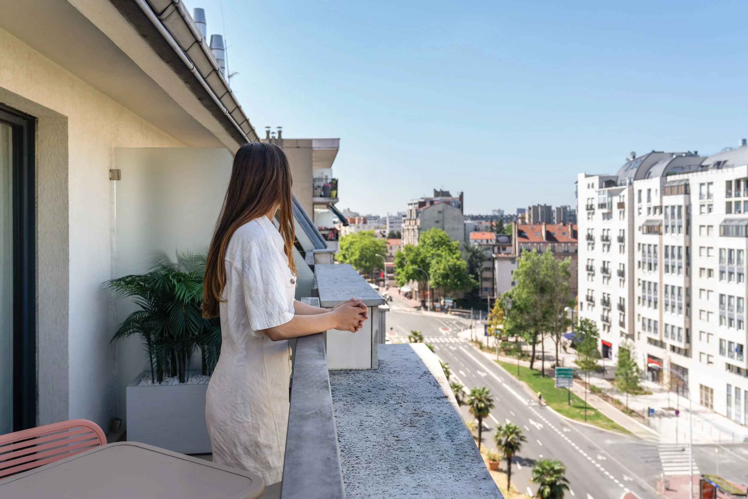 Une femme avec de longs cheveux bruns, portant une robe beige, se tient sur un balcon, regardant la ville avec des immeubles et des arbres verts sous un ciel clair et ensoleillé.