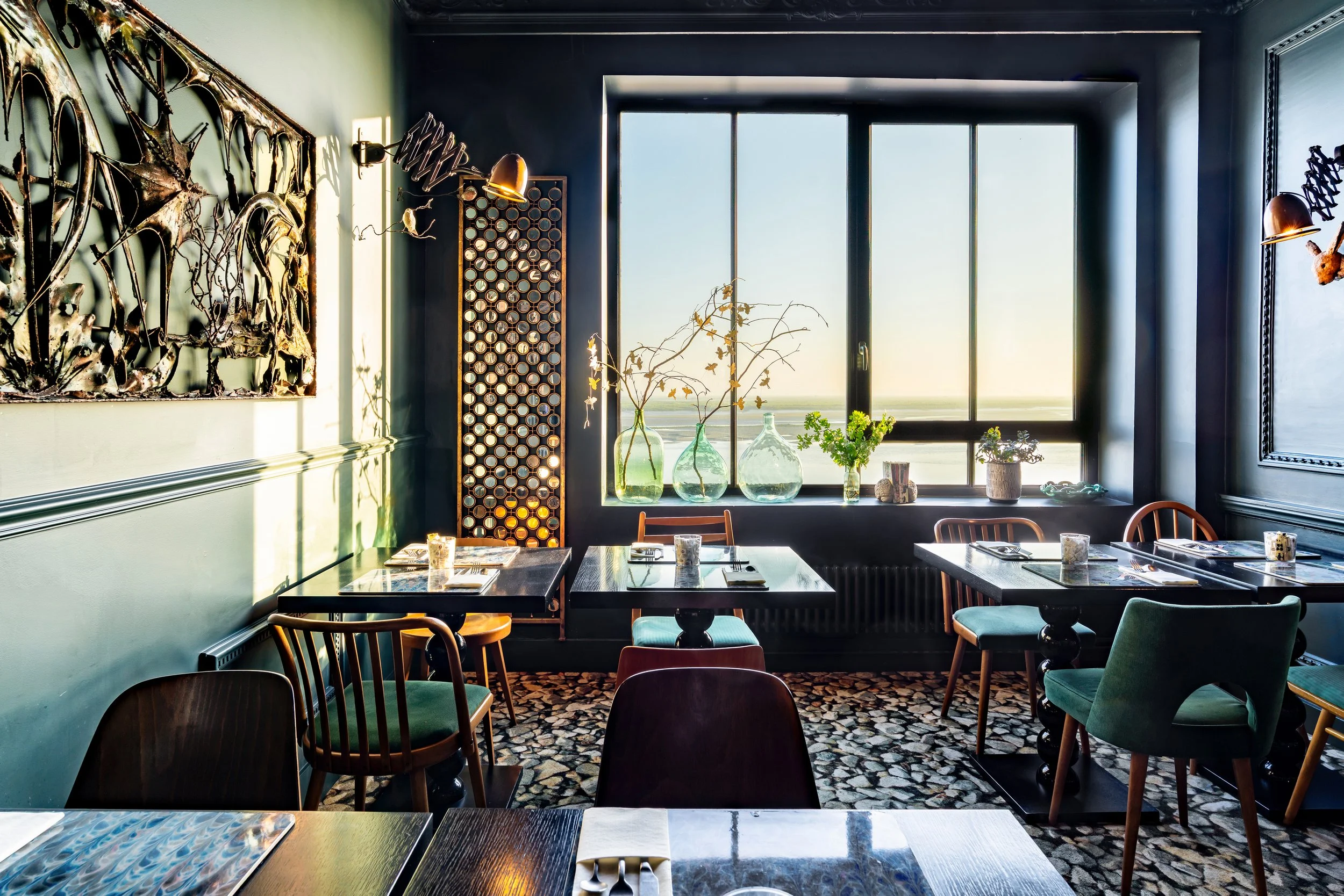 Salle à manger dans un restaurant avec un grand vitrage, des tables en bois noires, des chaises variées, des vases avec branches et plantes, décorations murales métalliques, et un sol en mosaïque, éclairée par la lumière naturelle