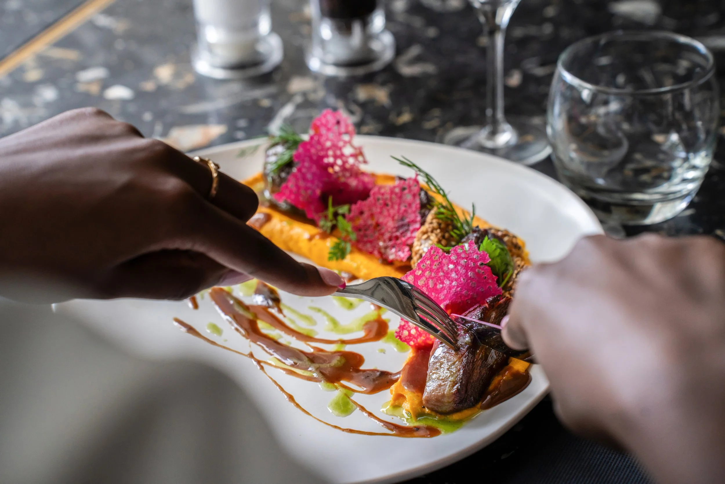 Une personne déguste un plat gourmet avec des légumes, des sauces colorées et des éléments décoratifs en rose sur une assiette blanche, au restaurant.
