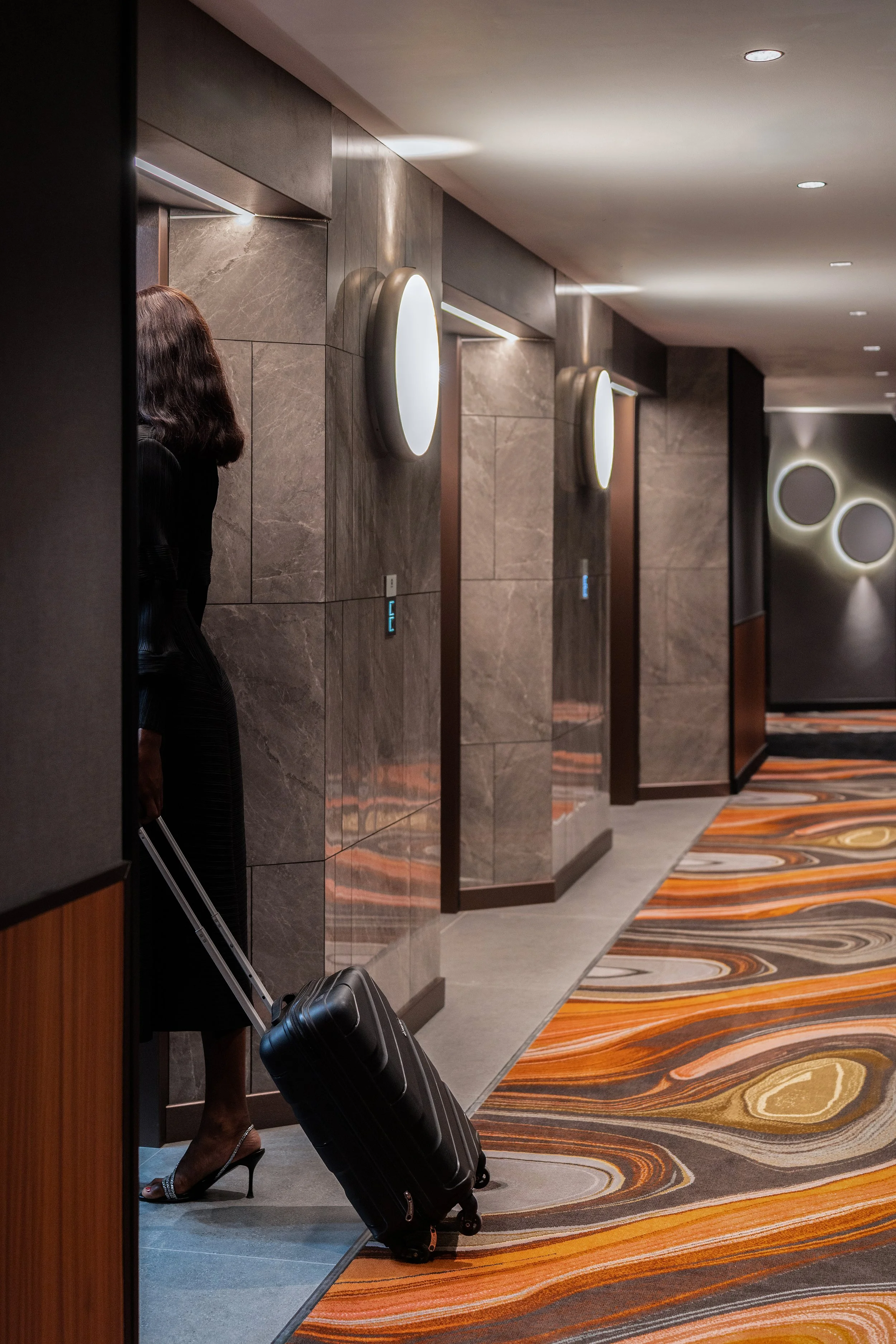 Une femme vêtue d'une robe noire et de talons hauts tire une valise noire dans un couloir d'hôtel avec des murs en marbre et un tapis rayé orange et gris.