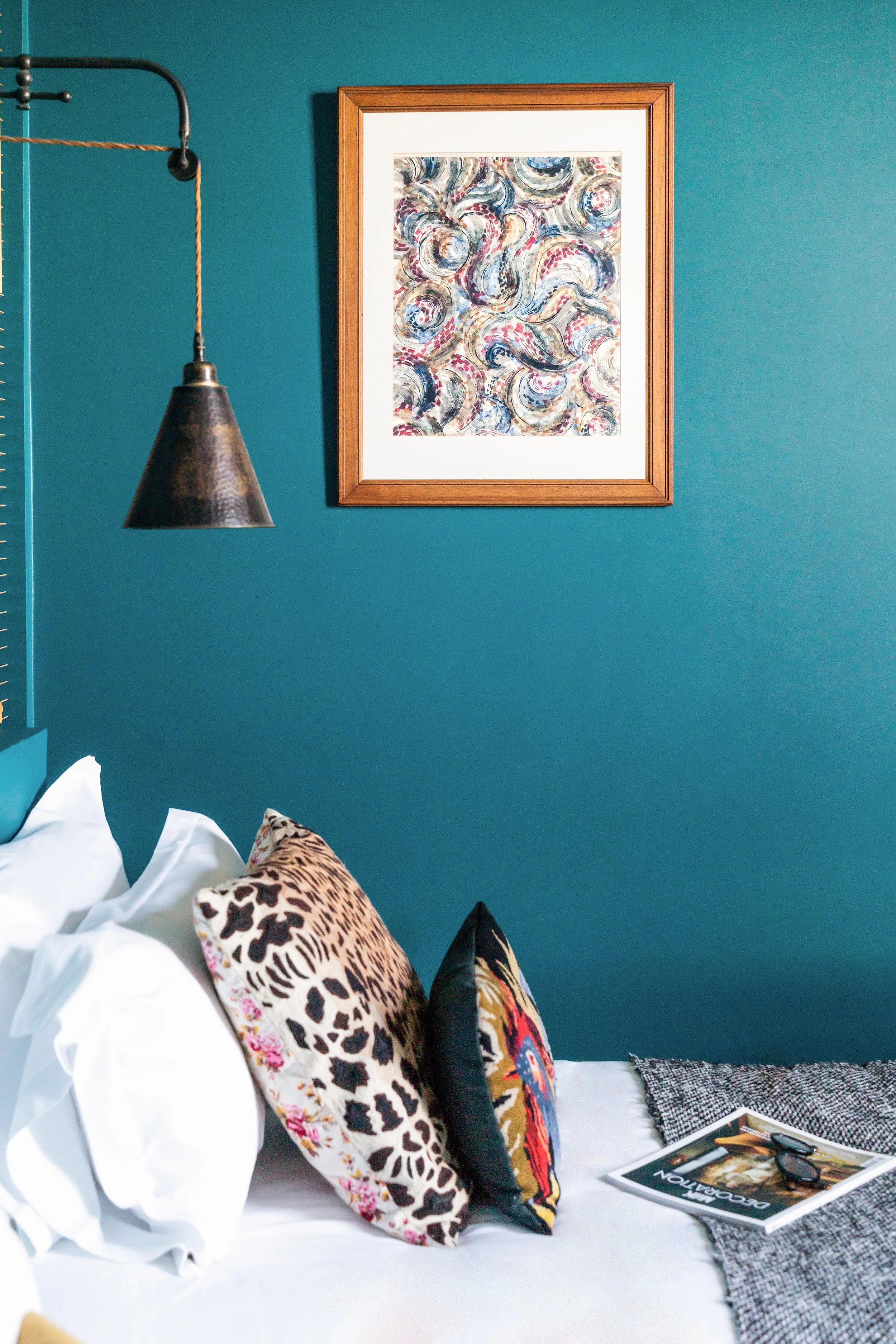 Mur bleu avec tableau abstrait, lampe suspendue en métal, lit avec des oreillers à motifs animaliers, magazine et lunettes sur la couverture du lit.