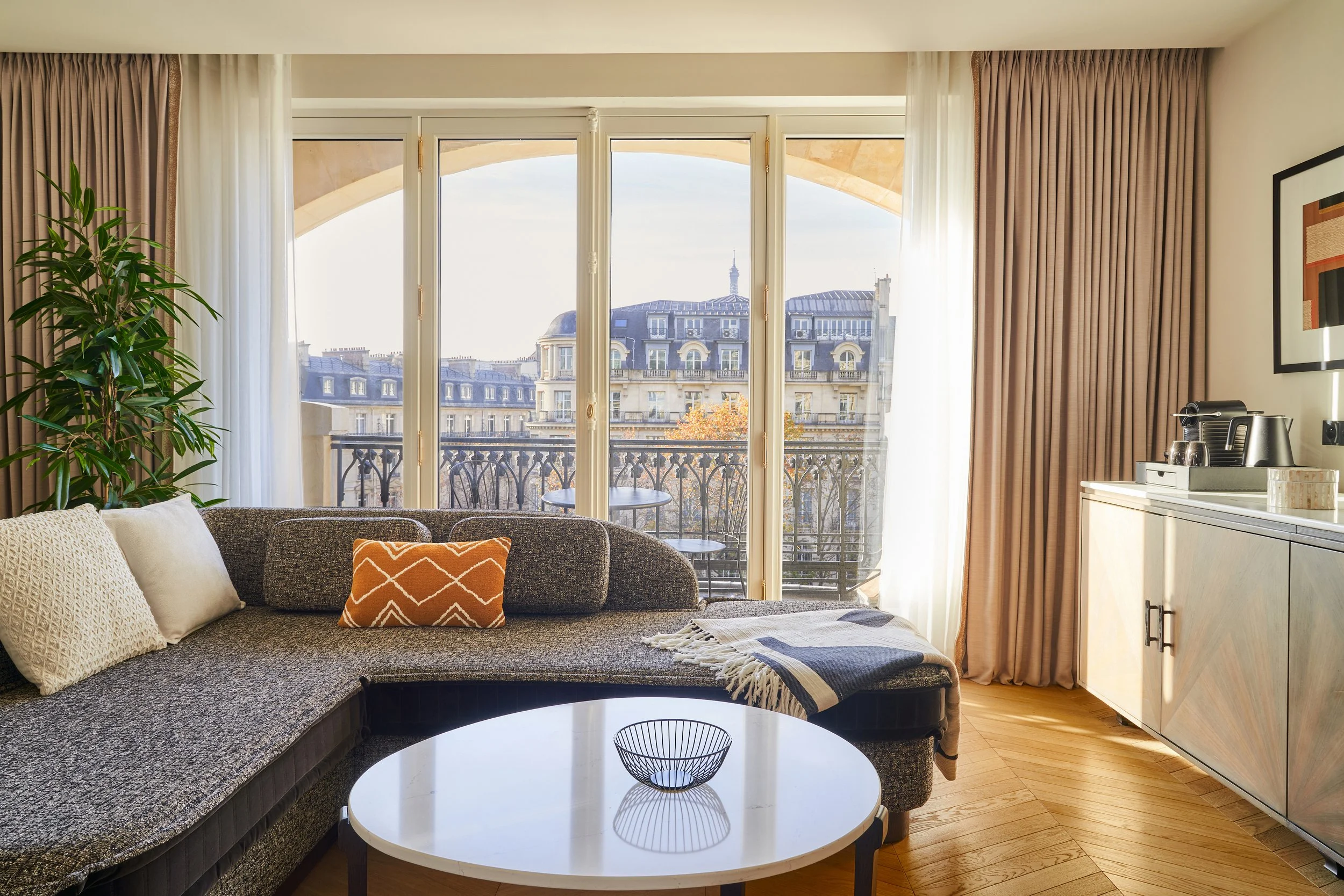 Salon avec canapé, table basse ronde, grande baie vitrée donnant sur un balcon avec vue sur des bâtiments haussmanniens parisien