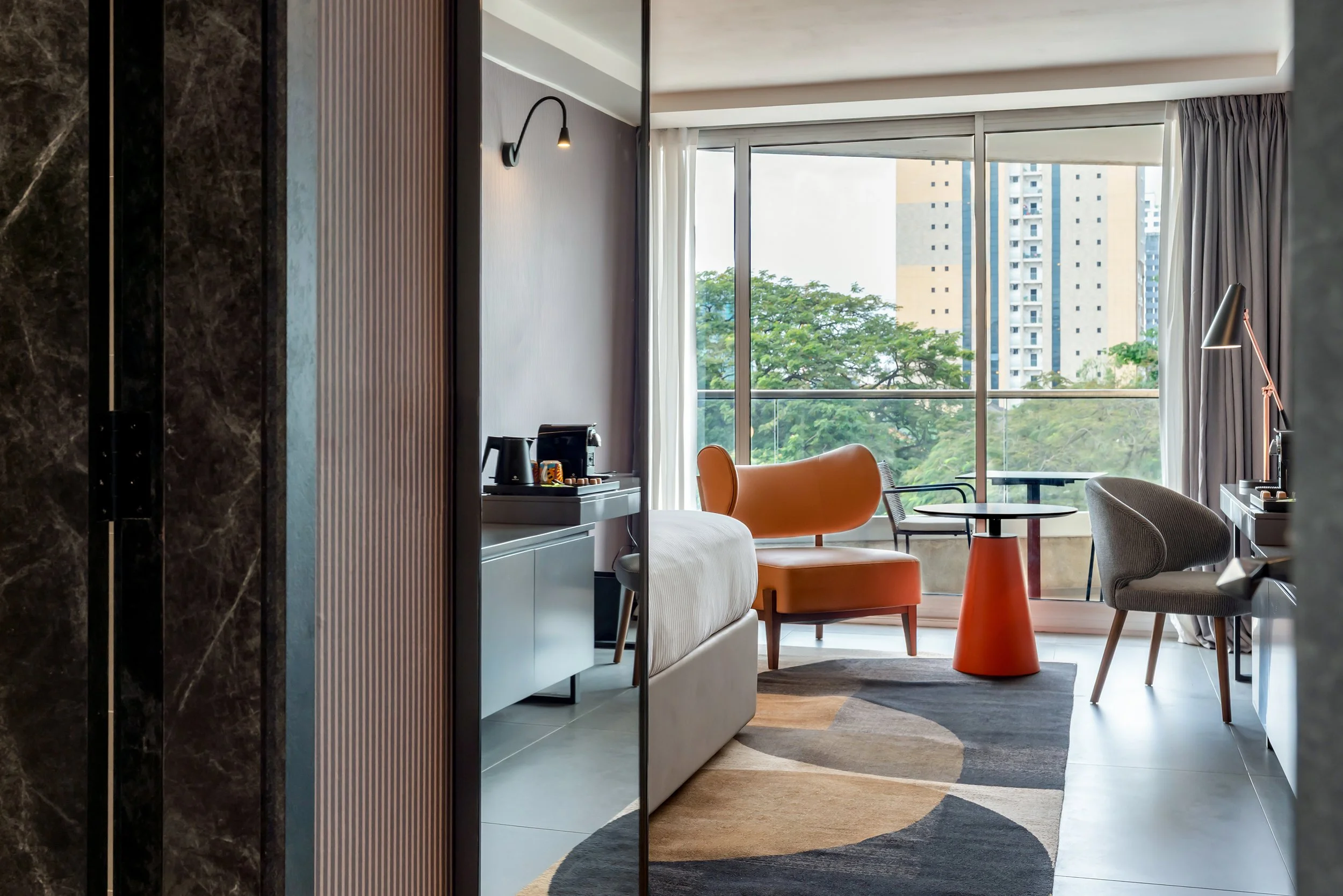 Intérieur moderne d'une chambre d'hôtel avec un coin salon, une table ronde, une lampe de bureau, un fauteuil orange, un fauteuil gris, vue sur un balcon avec des bâtiments et des arbres en arrière-plan.