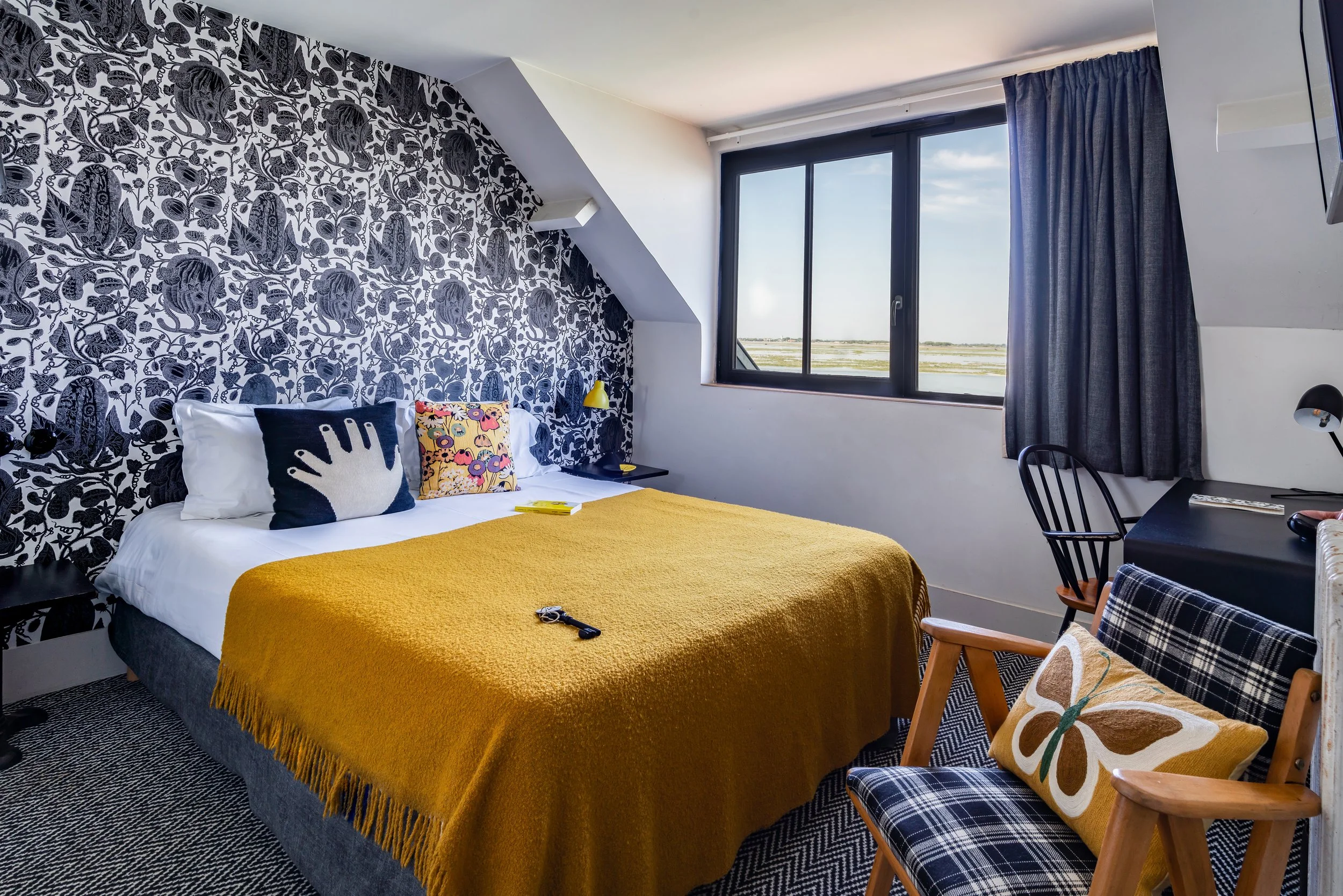 Chambre avec mur à motif floral en noir et blanc, lit avec une couverture jaune, coussins colorés, fenêtre avec vue de paysage, et fauteuil en bois avec coussin à motifs.