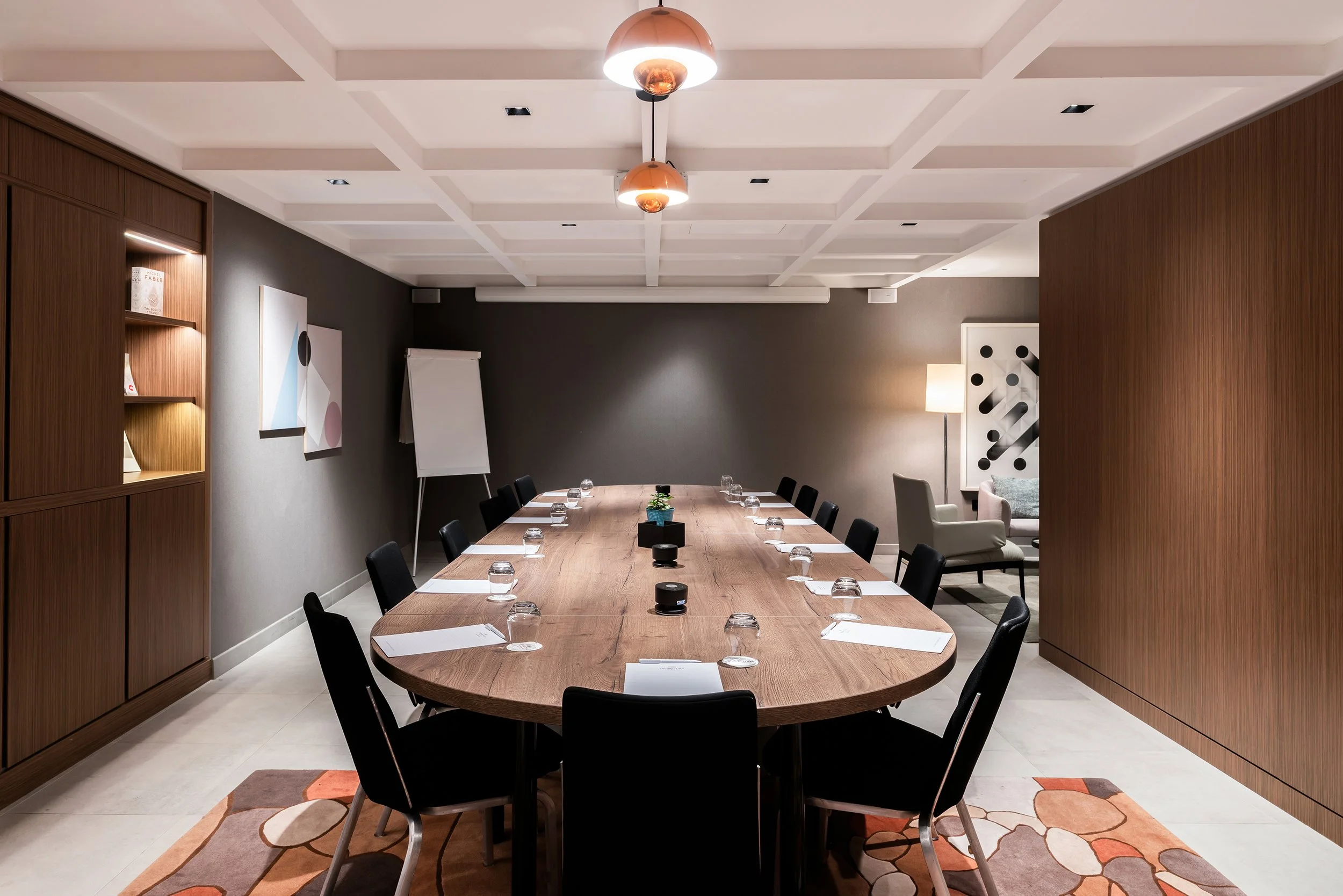 Salle de réunion boardroom avec une grande table ovale en bois, entourée de chaises noires, avec des feuilles et des verres d'eau, lampes au plafond, tableaux et autres meubles dans un décor moderne.