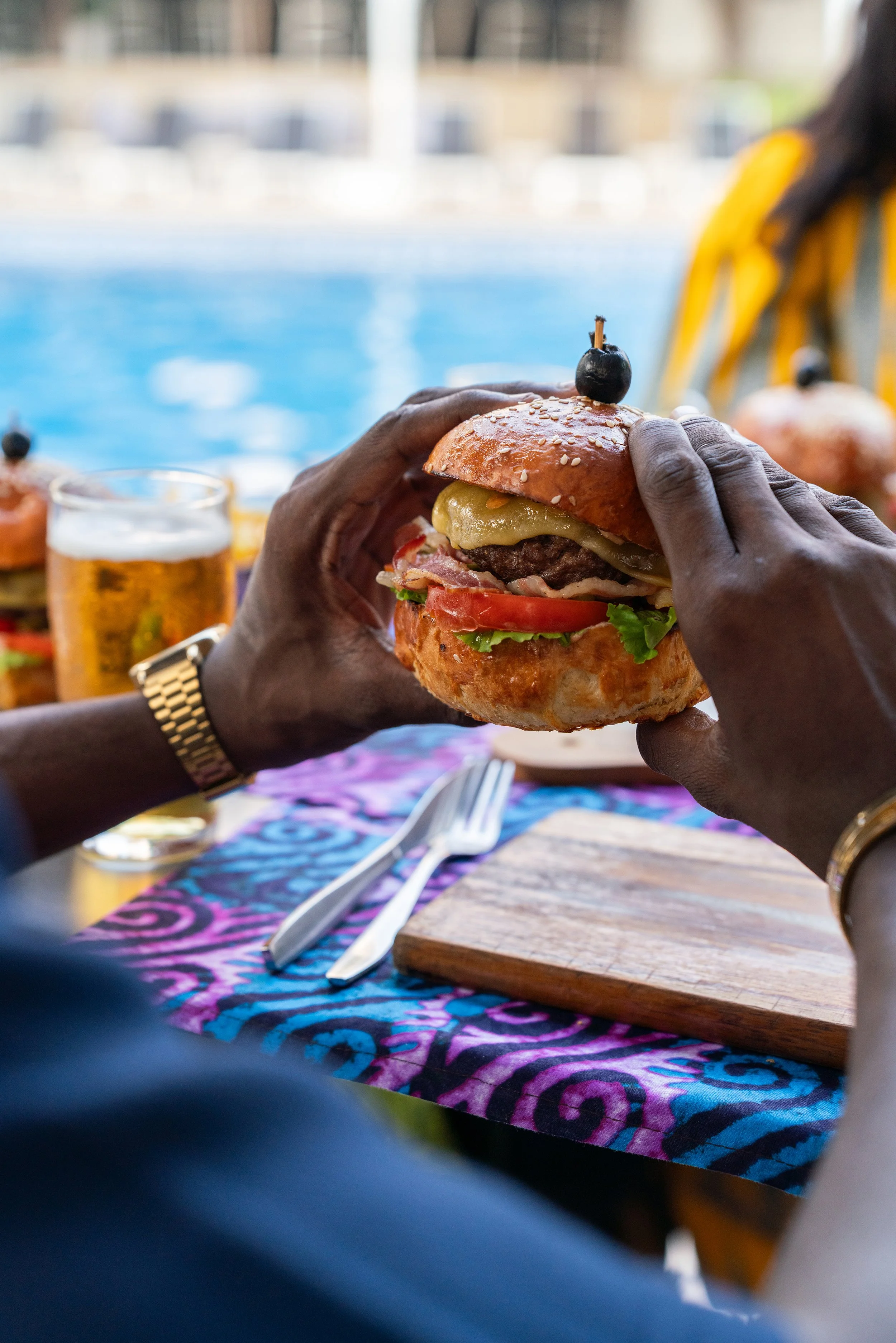 Une personne tient un burger avec des ingrédients visibles comme viande, tomate, laitue, fromage, et une olive en haut, avec une piscine en arrière-plan.