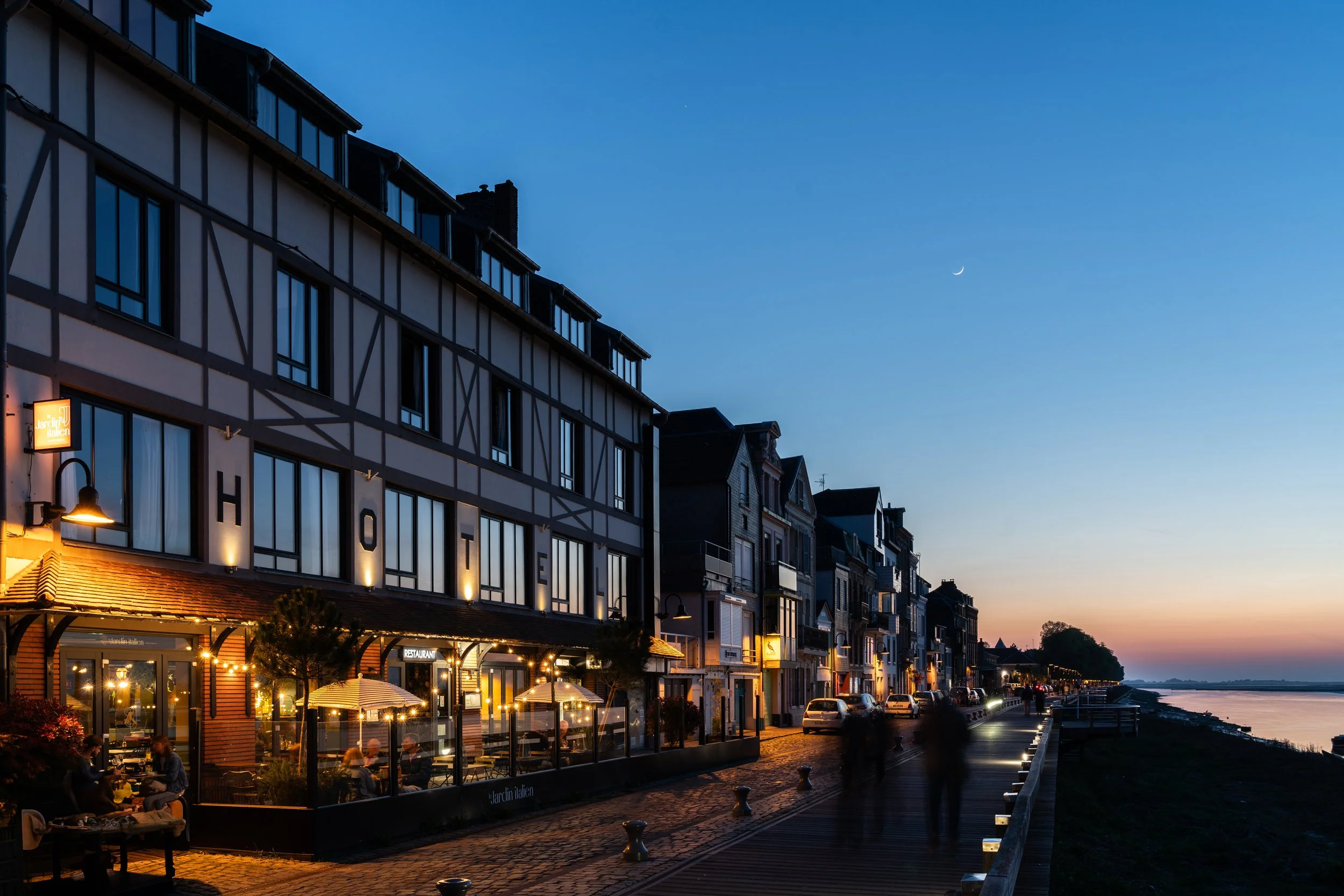 Rue bordée de bâtiments résidentiels et commerciaux éclairés au crépuscule, au bord d'une rivière, avec un ciel clair