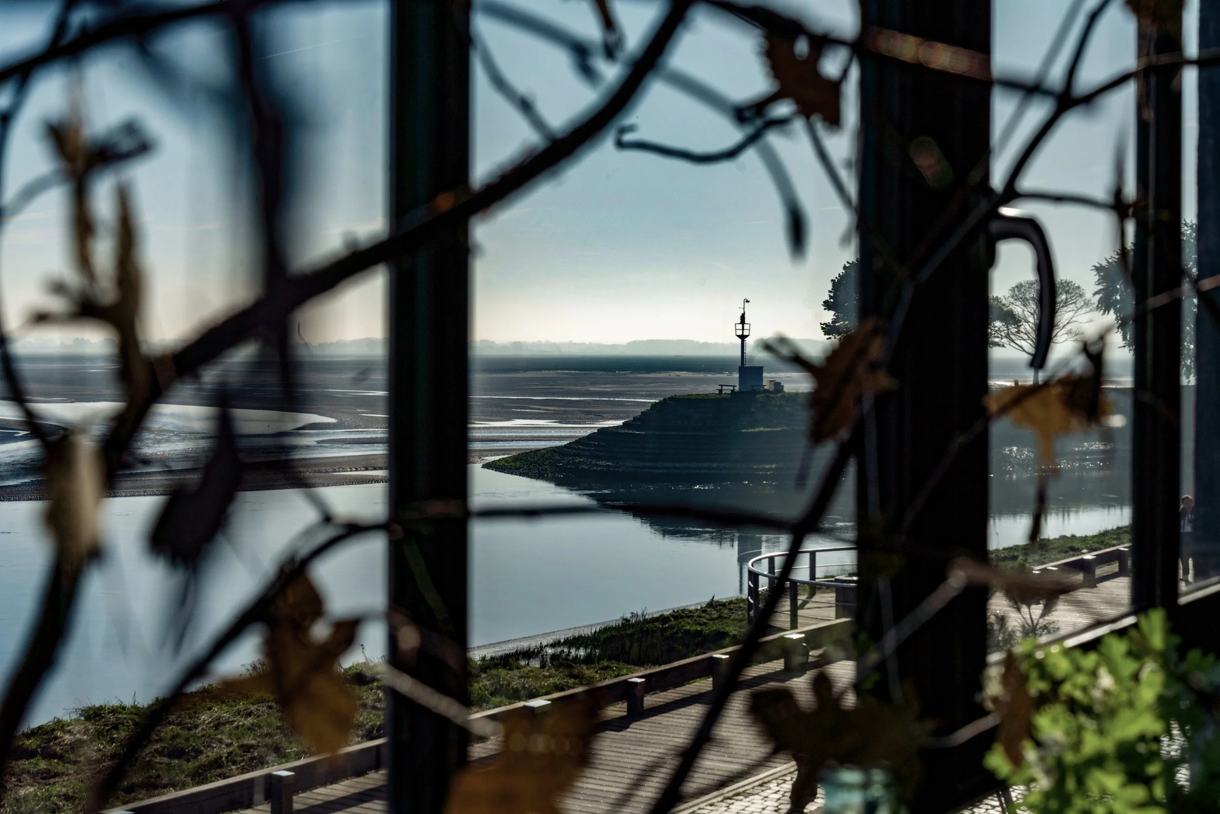 Vue d'un paysage avec un phare au loin, vue à travers une structure avec des branches et des feuilles sèches, reflet de l'eau et un chemin en bois.