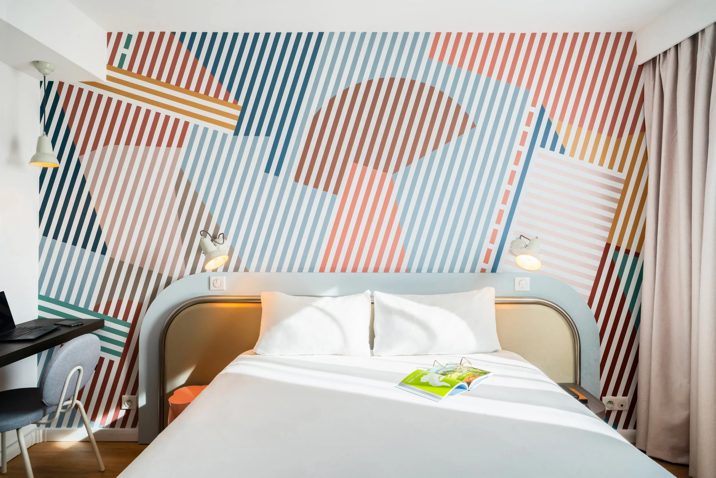 Chambre d'hôtel moderne avec mur en décoration géométrique colorée derrière un lit avec deux oreillers blancs. Il y a une lampe de lecture à chaque côté du lit et un livre ouvert sur la couverture verte sur le lit. À gauche, un coin avec un bureau, u