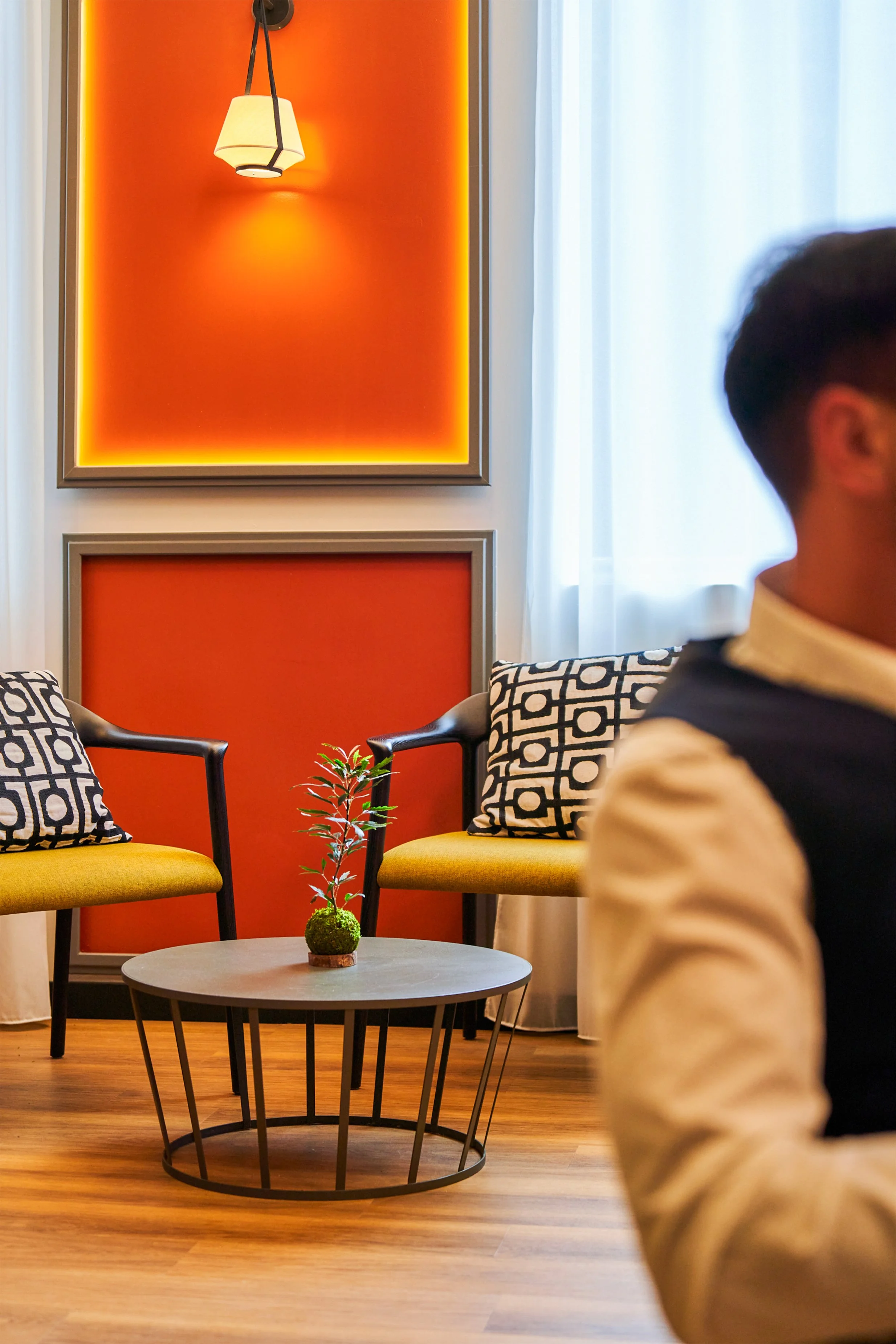 Intérieur moderne avec deux fauteuils jaunes, coussins à motifs noirs et blancs, une petite table ronde avec une plante verte, murs orange avec accents lumineux et une fenêtre avec rideaux blancs.