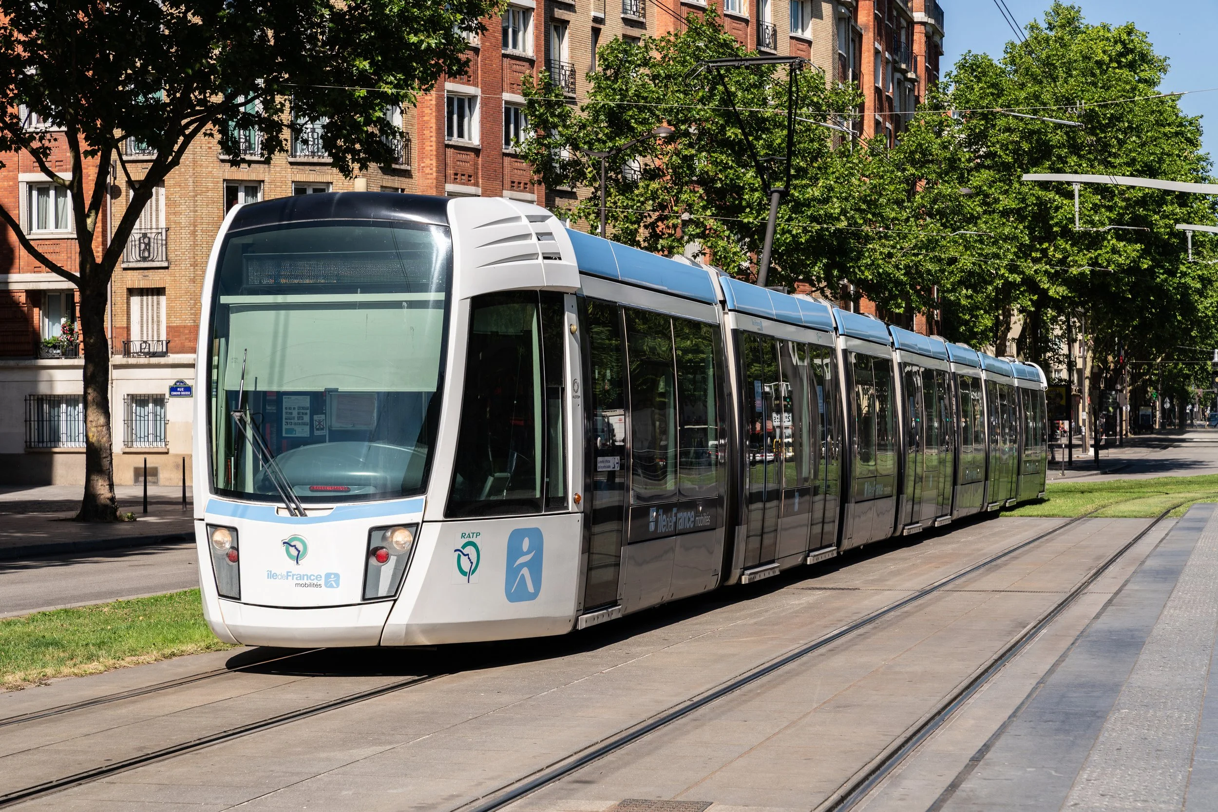 Un tramway moderne blanc et noir circulant sur des rails en ville, entouré d'arbres et de bâtiments résidentiels en arrière-plan.