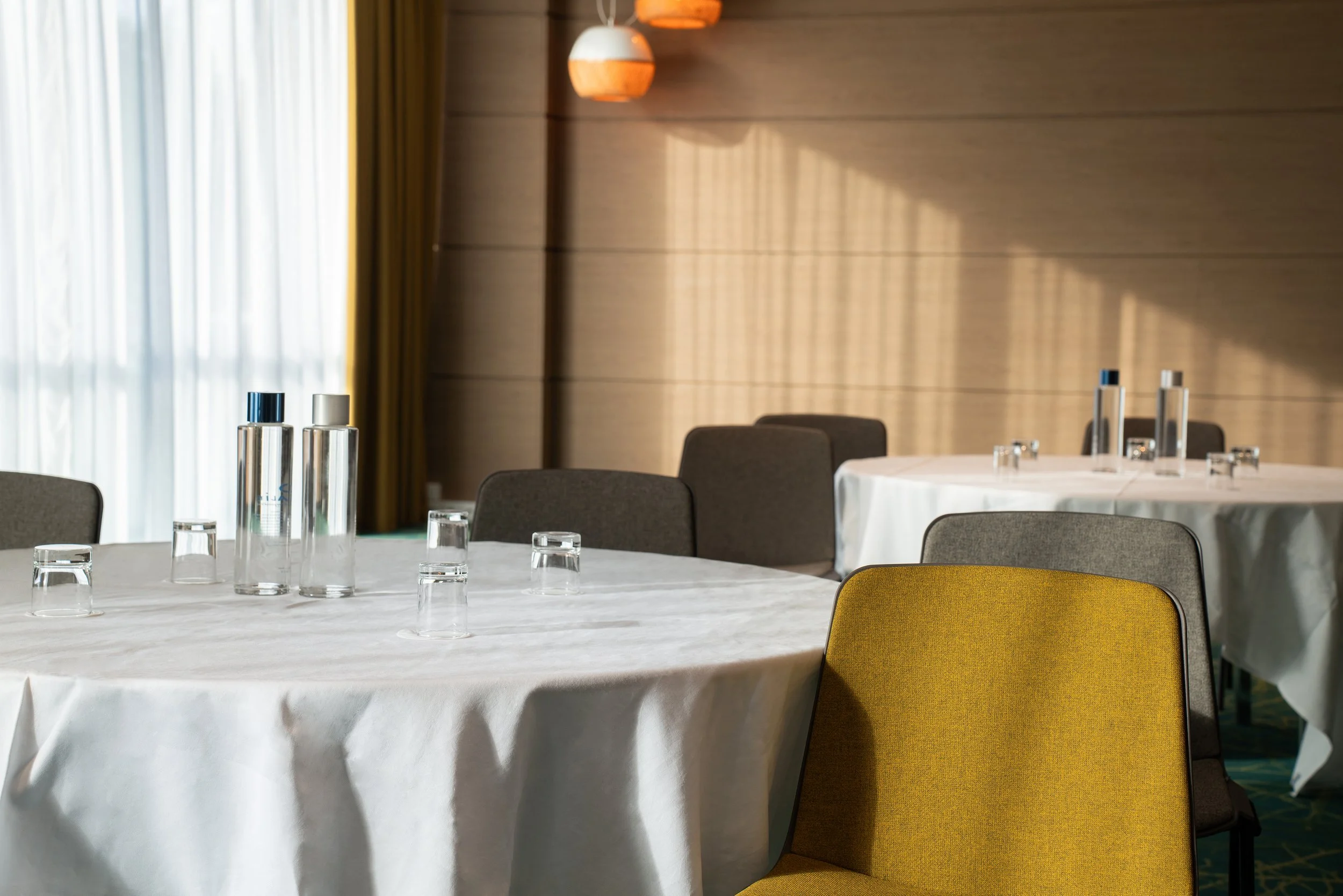 Salle de réunion avec tables rondes, verres, bouteilles d'eau et chaises en tissu de différentes couleurs