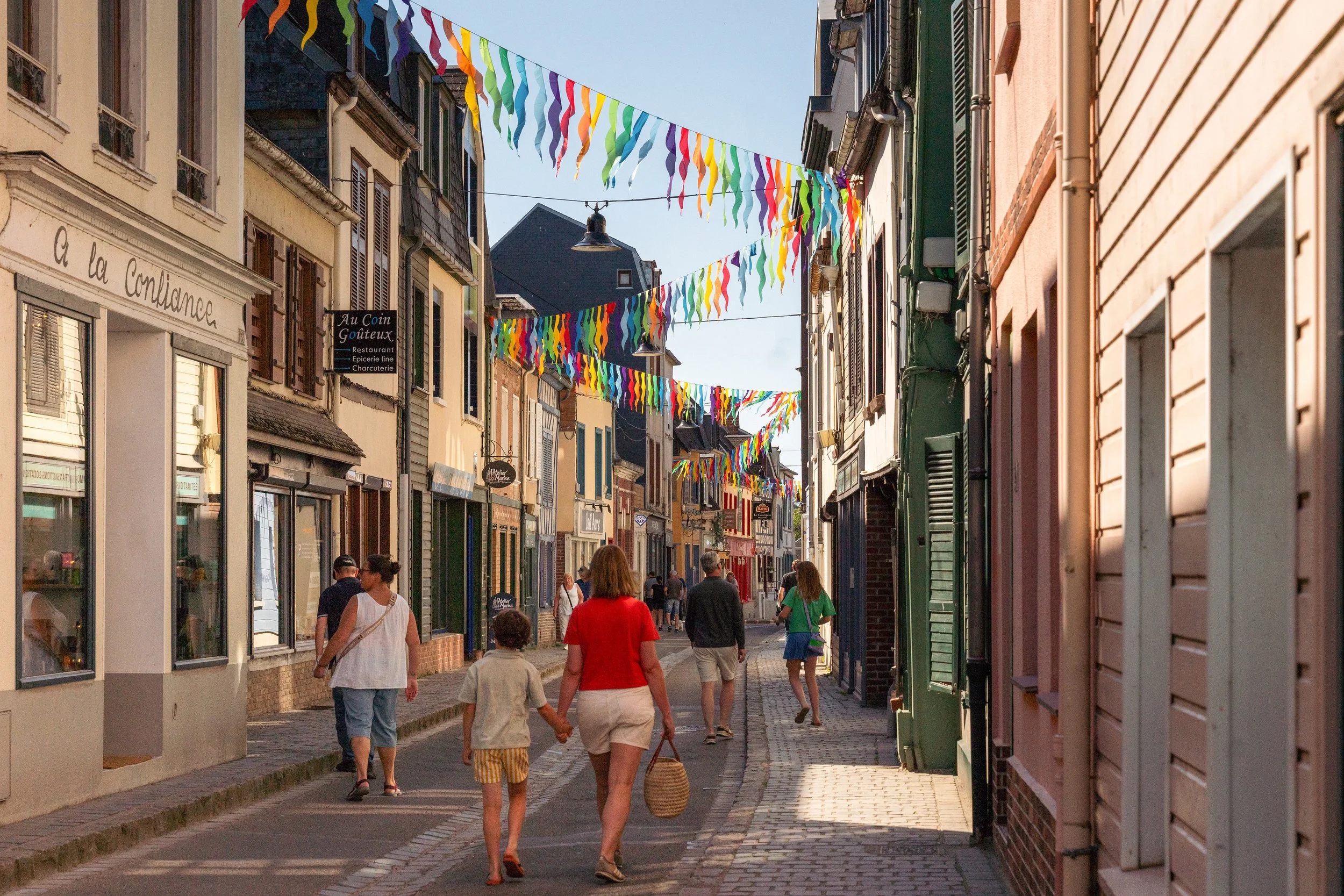 Une rue pavée dans une ville avec des boutiques colorées et des décorations de fanions multicolores suspendus au-dessus de la rue, avec des personnes marchant et quelques enfants en promenade.