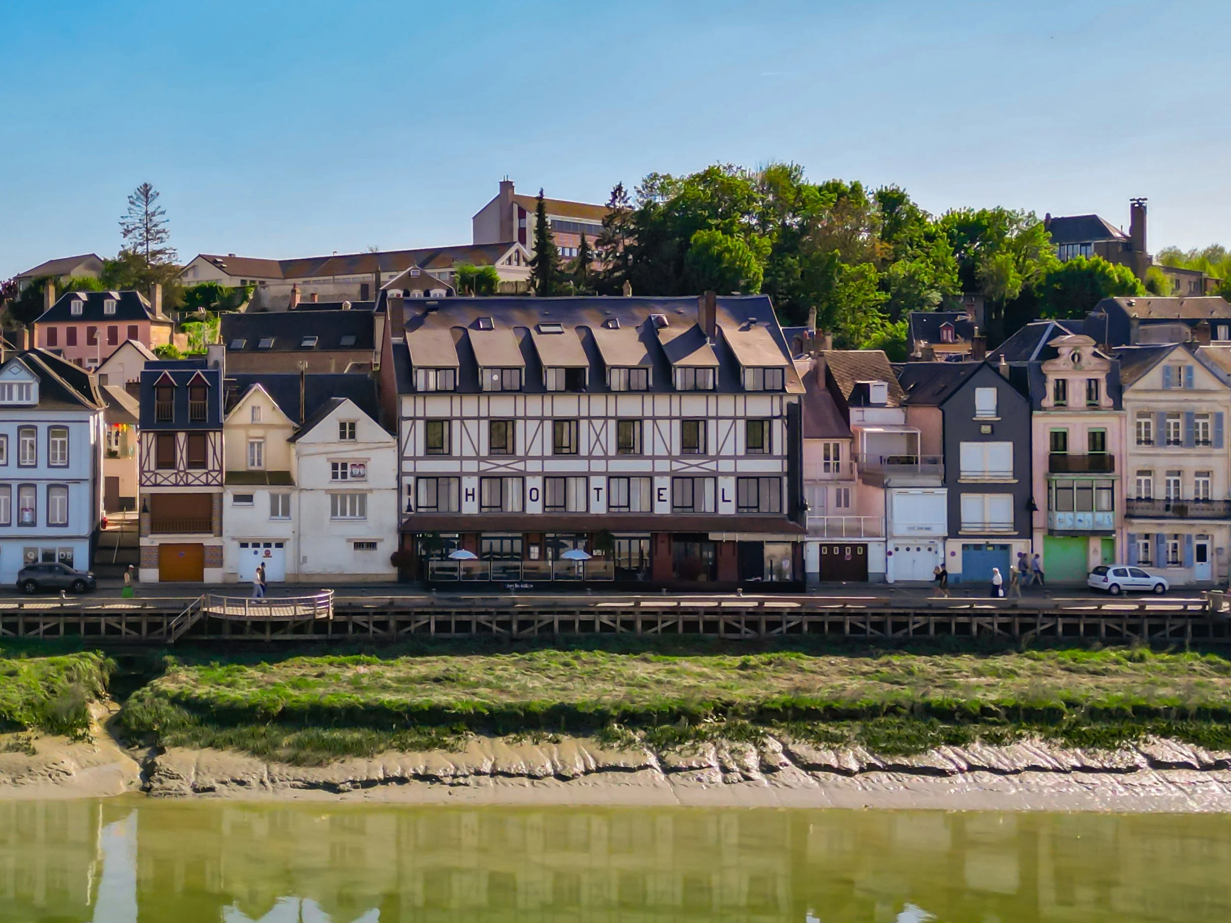 Maisons colorées avec un hôtel au centre, le long de la rivière, sous un ciel bleu, avec des arbres en arrière-plan.