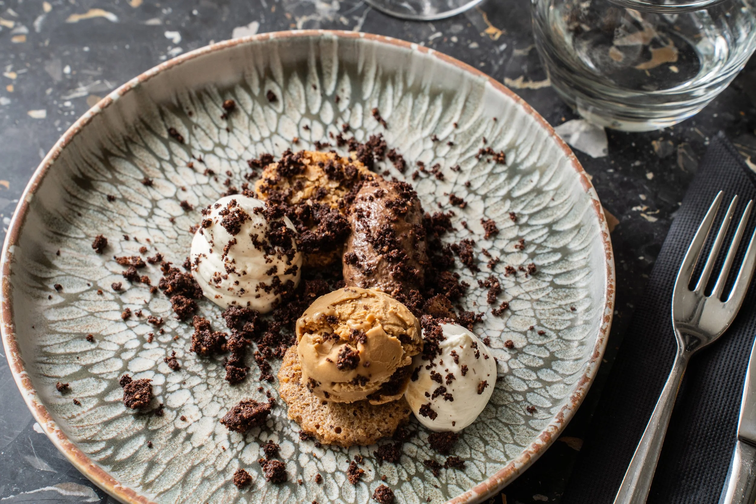 Dessert avec plusieurs boules de glace chocolat, caramel et vanille, garnies de miettes de biscuits, servi sur une assiette décorative, avec une fourchette, un verre d'eau, et un napperon noir.