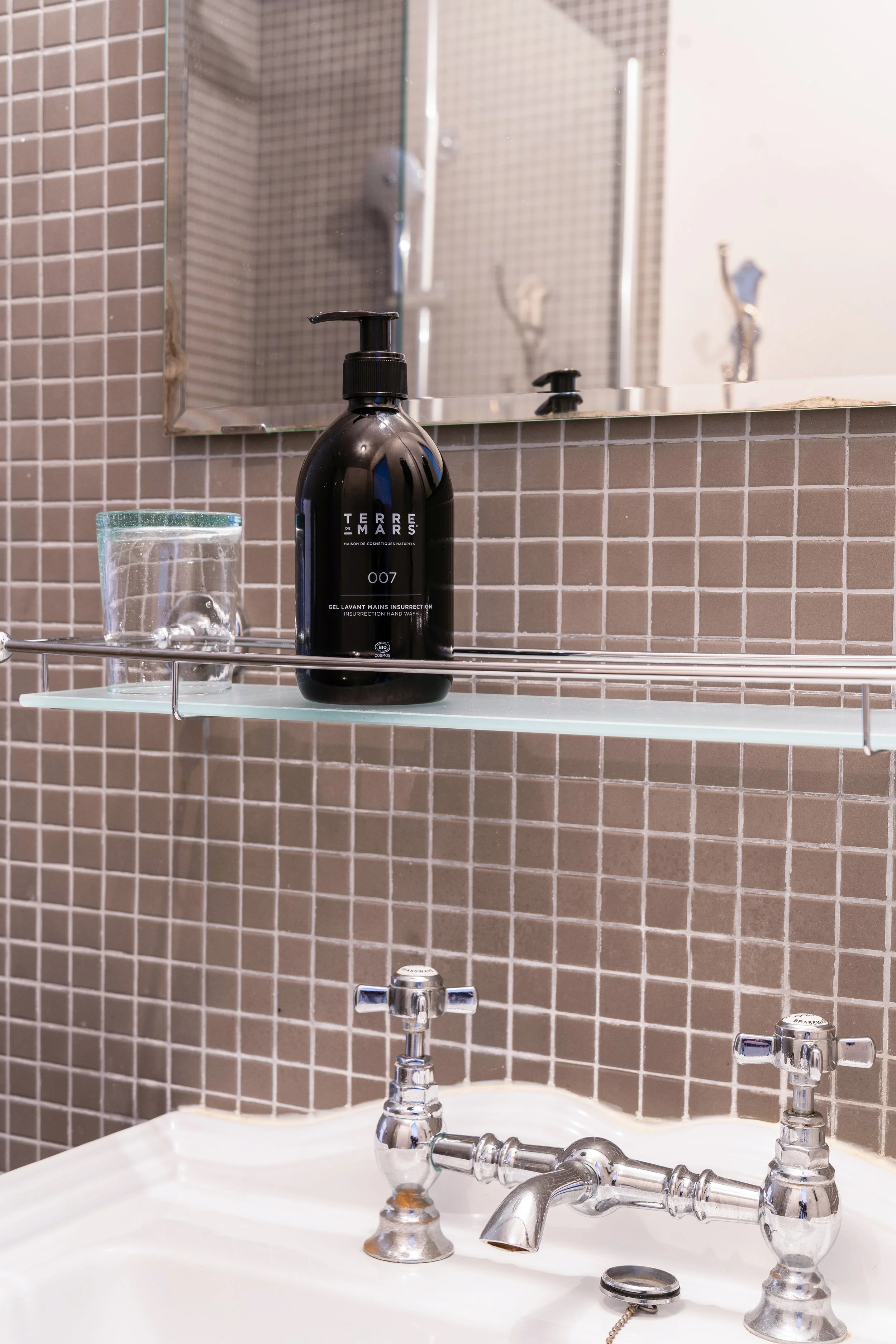 Étagère en verre avec un flacon de savon noir et un verre d'eau, miroir au mur, lavabo en céramique avec robinets en chrome dans une salle de bain.