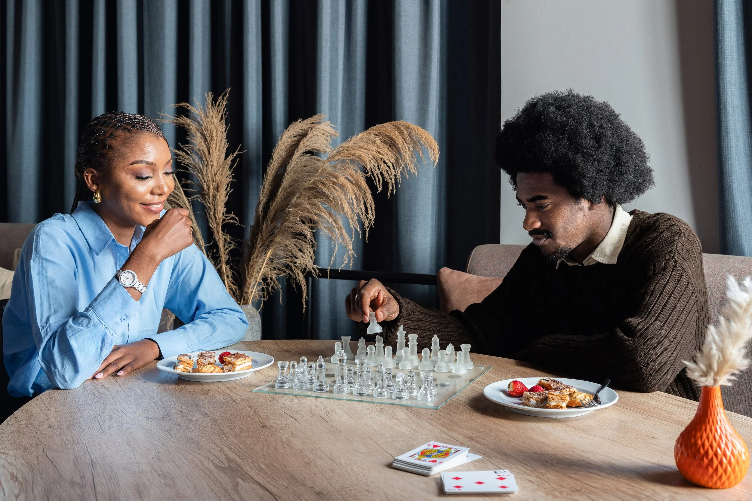 Une femme et un homme jouent aux échecs lors d'une pause détente dans le café d'un hôtel, avec des plats de desserts et un jeu de cartes sur la table.