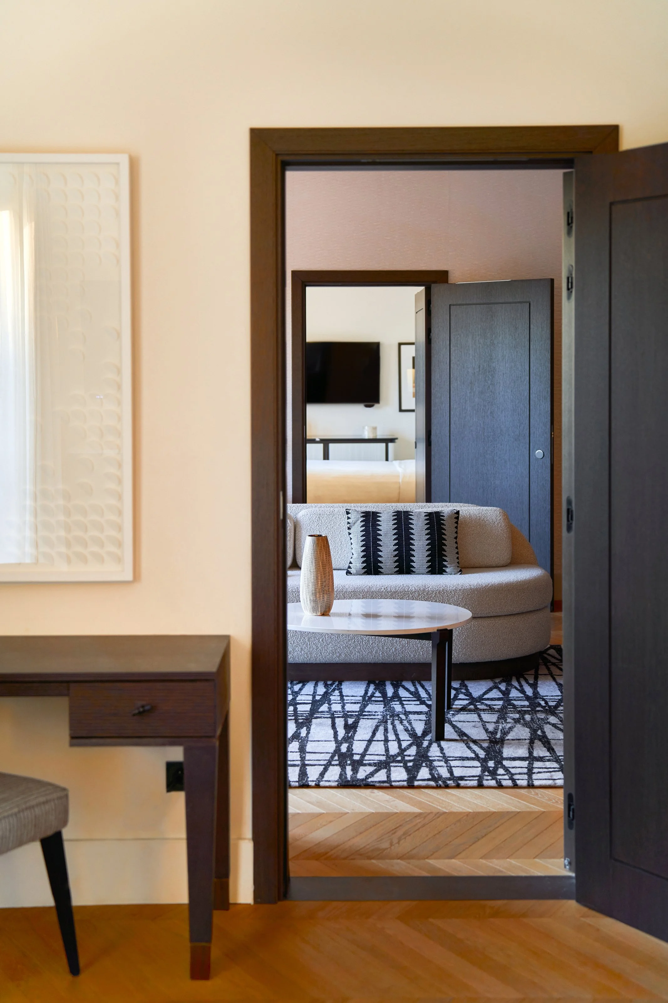 Vue à travers une porte ouverte montrant un salon avec un canapé beige, un coussin noir et blanc, une table basse avec une vase beige, un tapis noir et blanc, et un mur avec une télévision