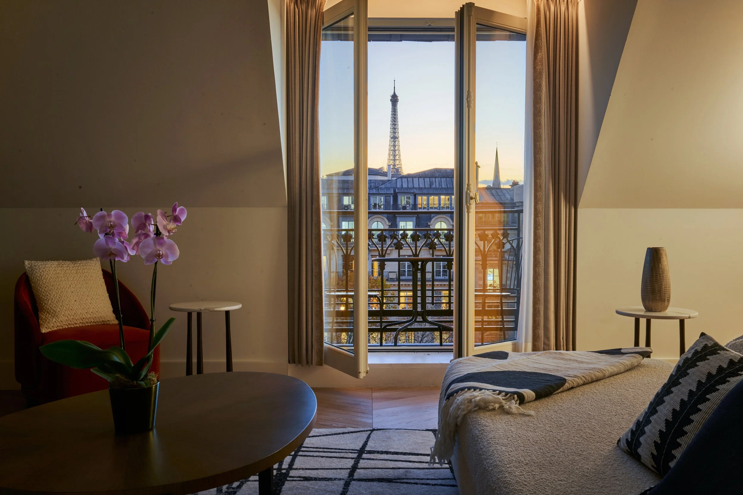 Salon intérieur avec lit, table basse, fauteuil orange, vase avec orchidée, vue sur la Tour Eiffel depuis un balcon pendant le couché de soleil.