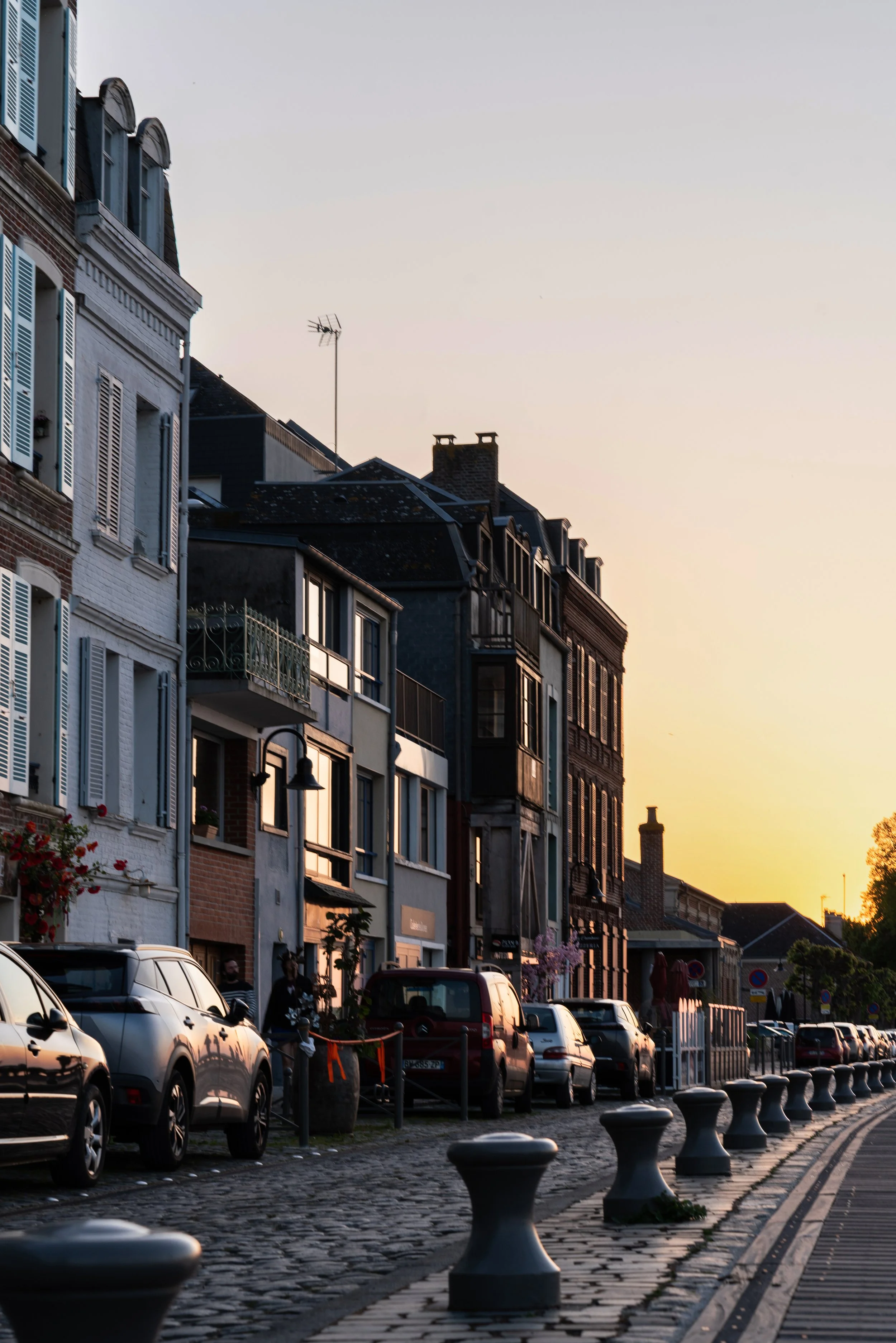 Rues pavée avec des voitures garées et bâtiments résidentiels sous un ciel au coucher du soleil.