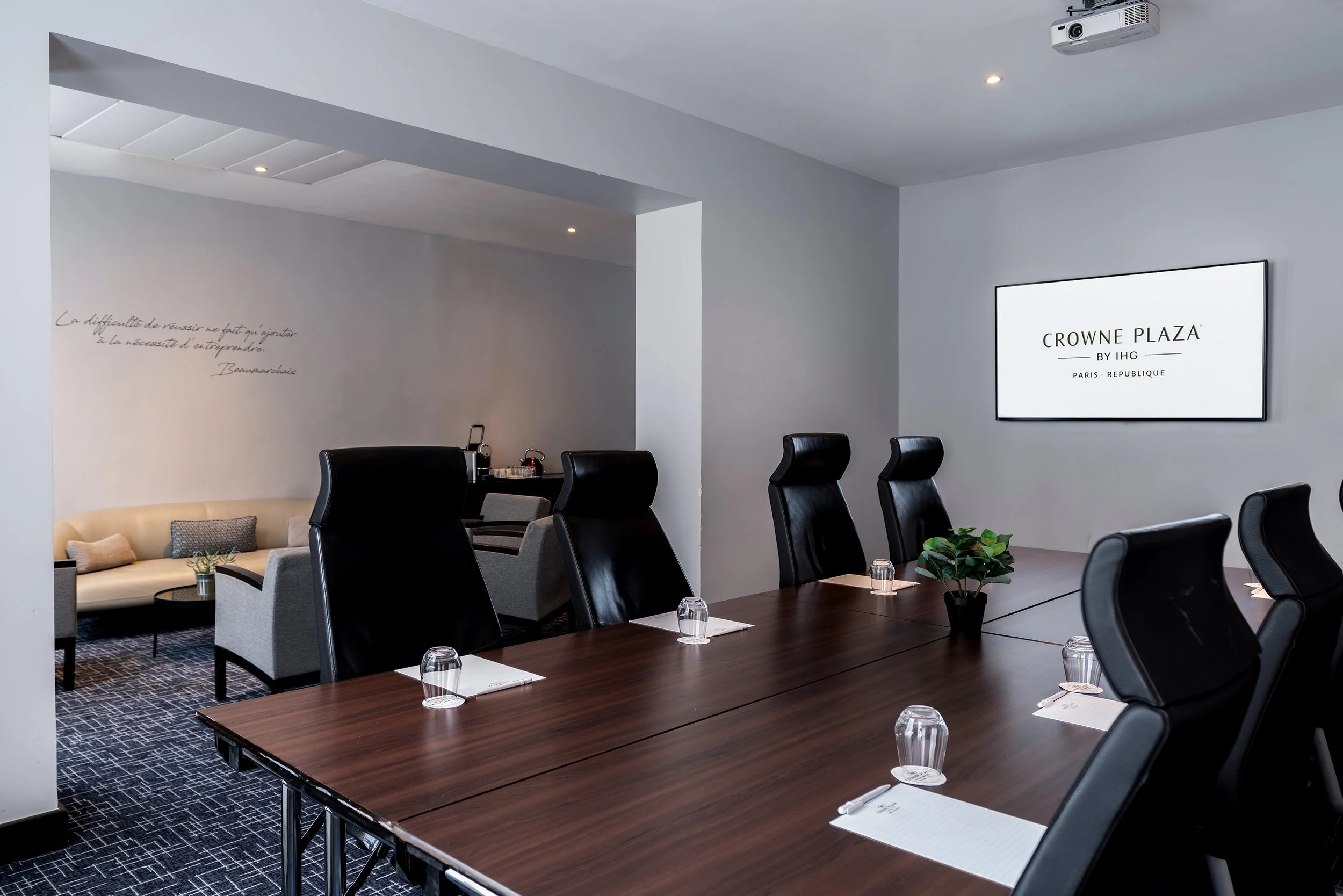 Salle de réunion moderne avec une grande table en bois, des chaises en cuir noir, des verres, des blocs-notes, une plante verte et un écran numérique affichant « Crowne Plaza, Paris République ».