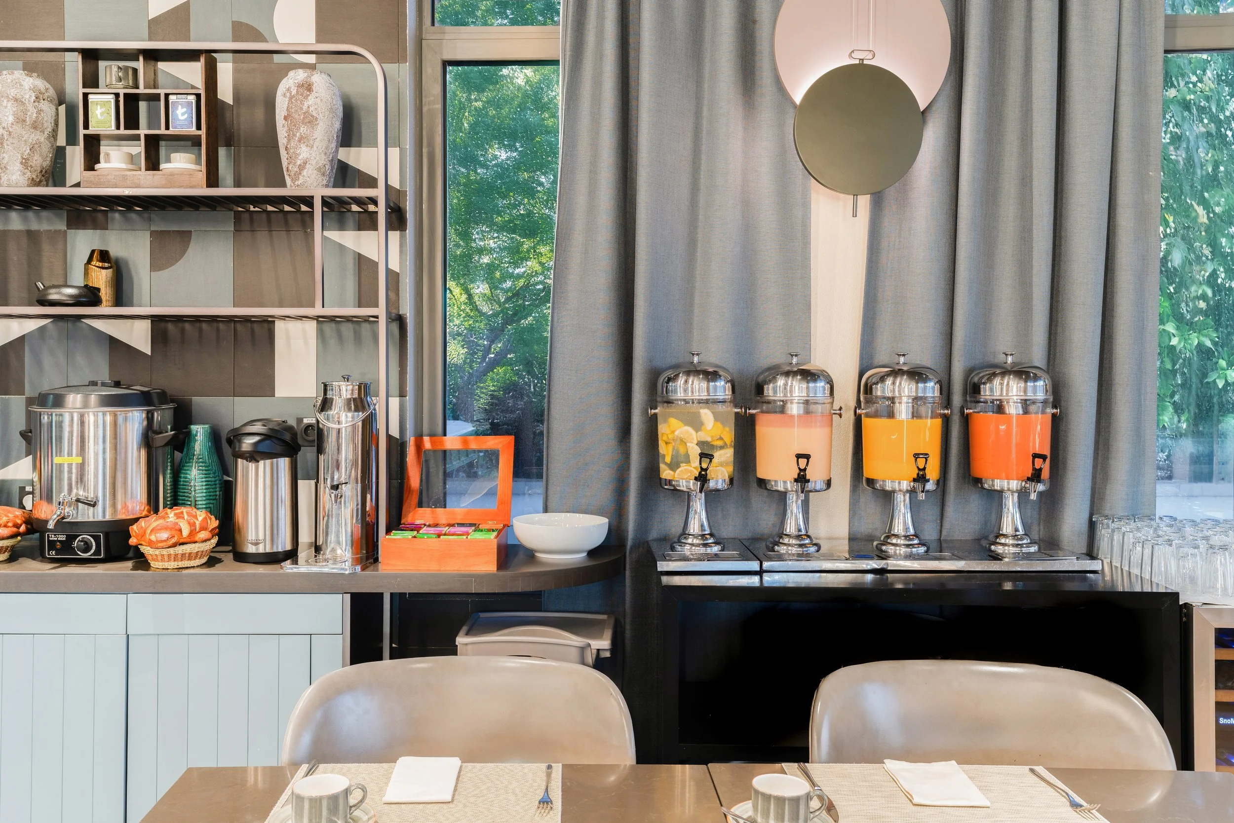 Station de boissons avec quatre distributeurs de jus à l'arrière-plan, vaisselle, théières et tasses sur la table avant, grande fenêtre avec rideaux gris en arrière-plan.