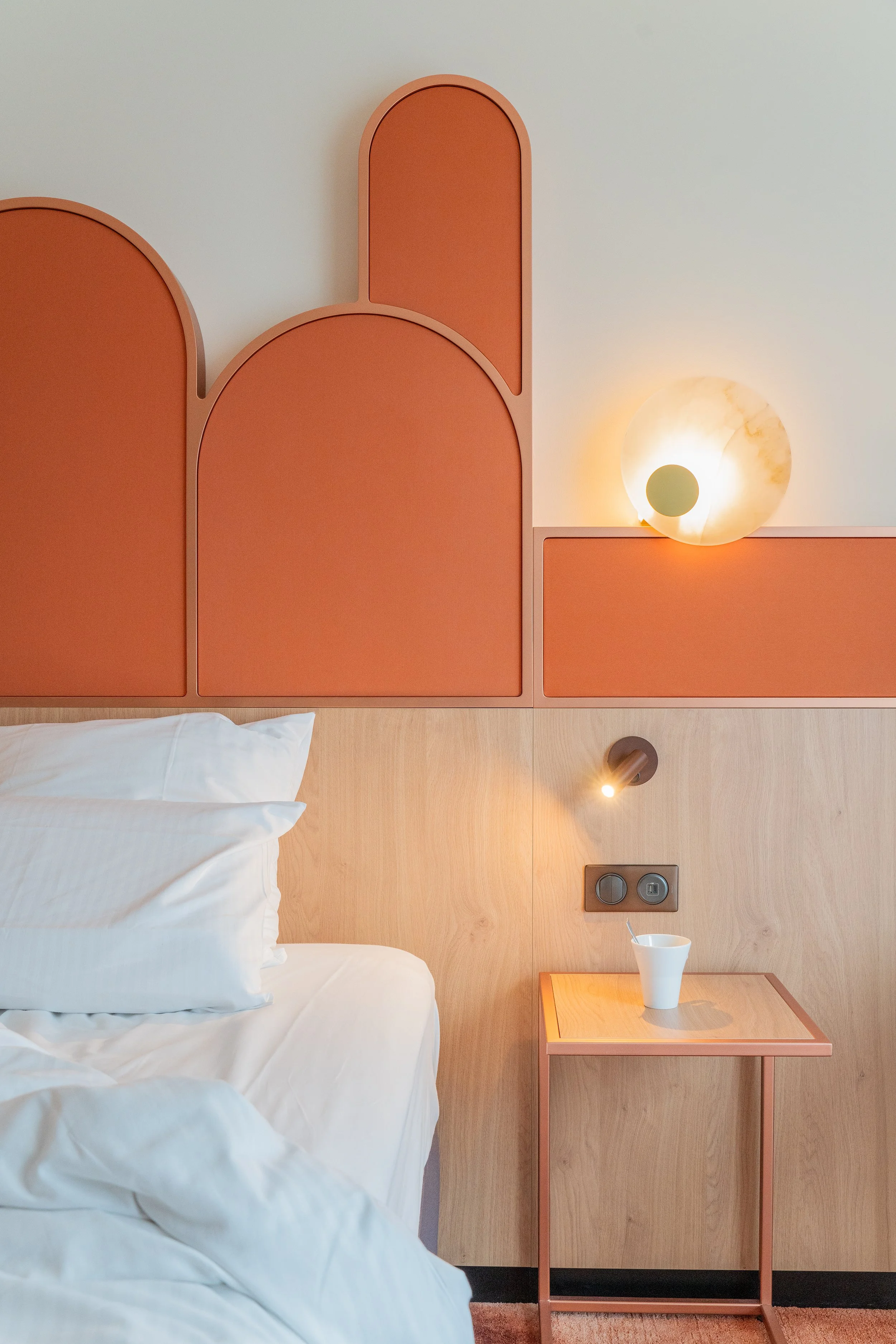 Chambre avec lit, tête de lit en bois clair, éléments de décoration murale orange, lampe de chevet en marbre, petite table en bois avec une tasse, prise électrique visible.