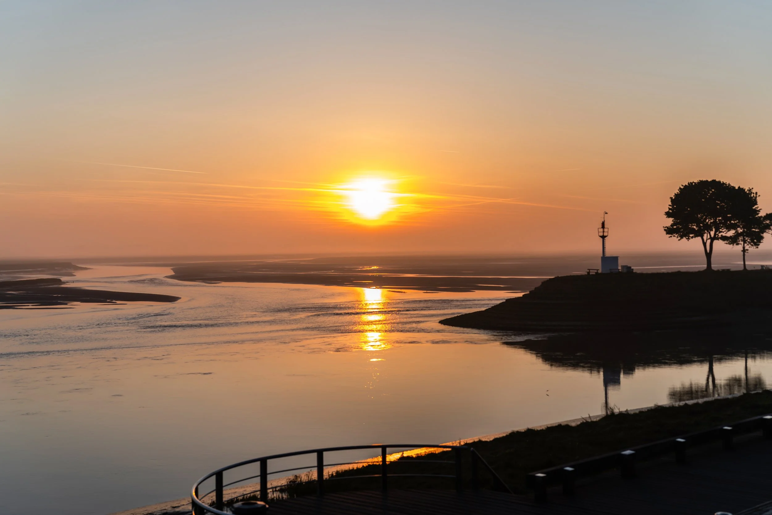 Coucher de soleil sur un fleuve avec un phare, deux arbres et un ciel coloré en arrière-plan.