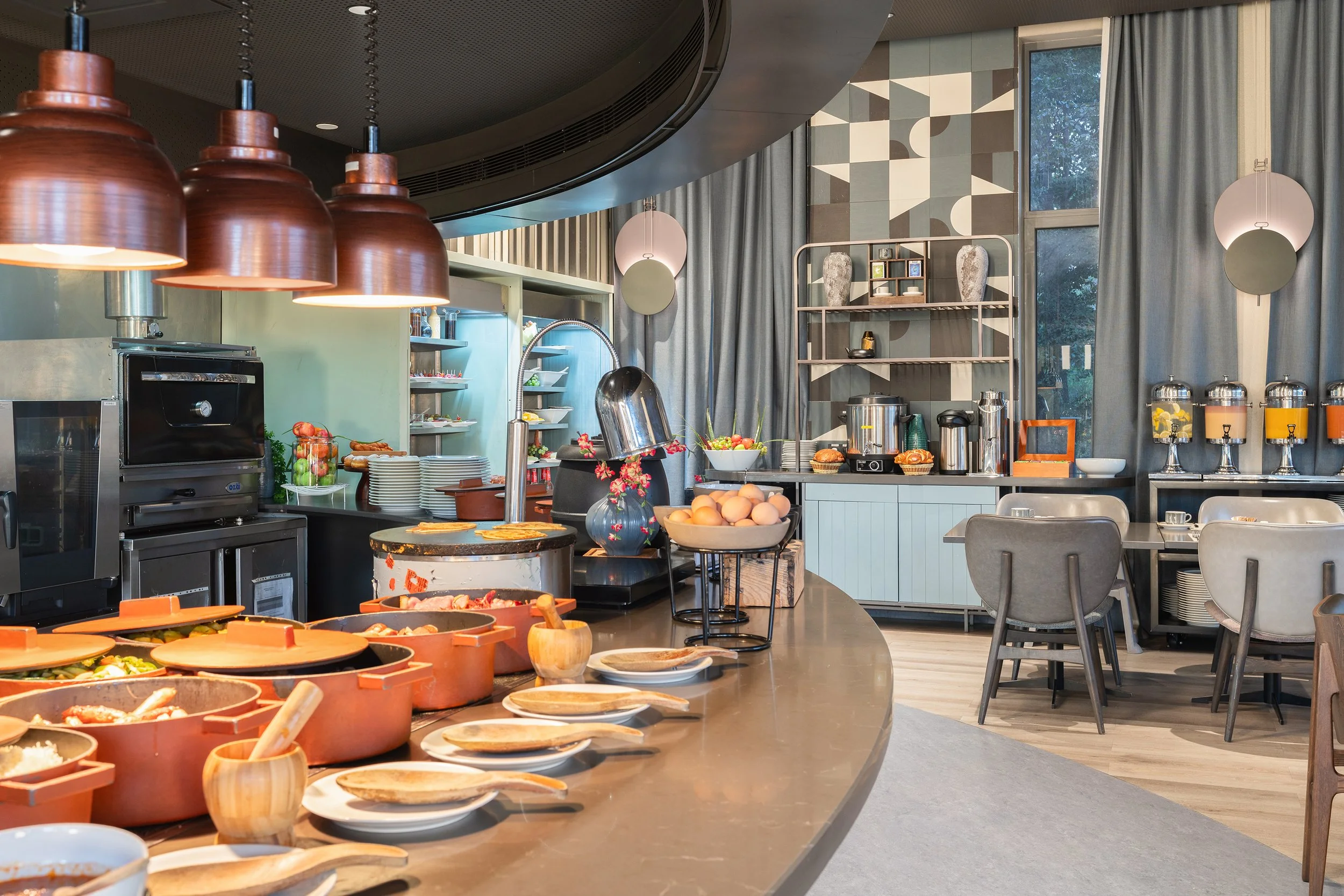 Buffet de petit-déjeuner avec plusieurs plats chauds, fruits, œufs, céréales, et ustensiles dans une salle lumineuse avec grande fenêtre et décoration moderne.