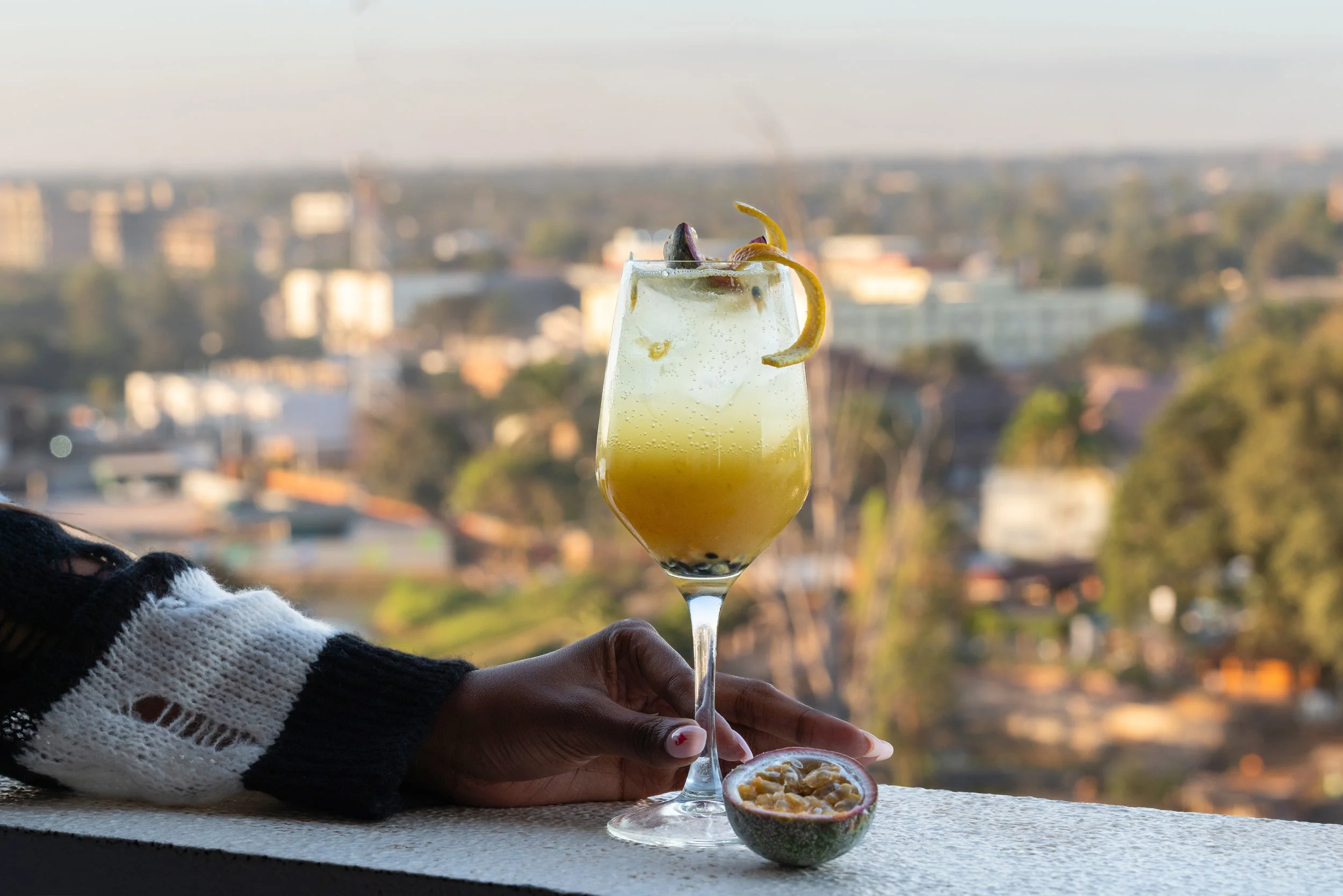 Verre de cocktail jaune avec une tranche de citron, posé sur un rebord avec un fond flou d'une ville et de la nature.