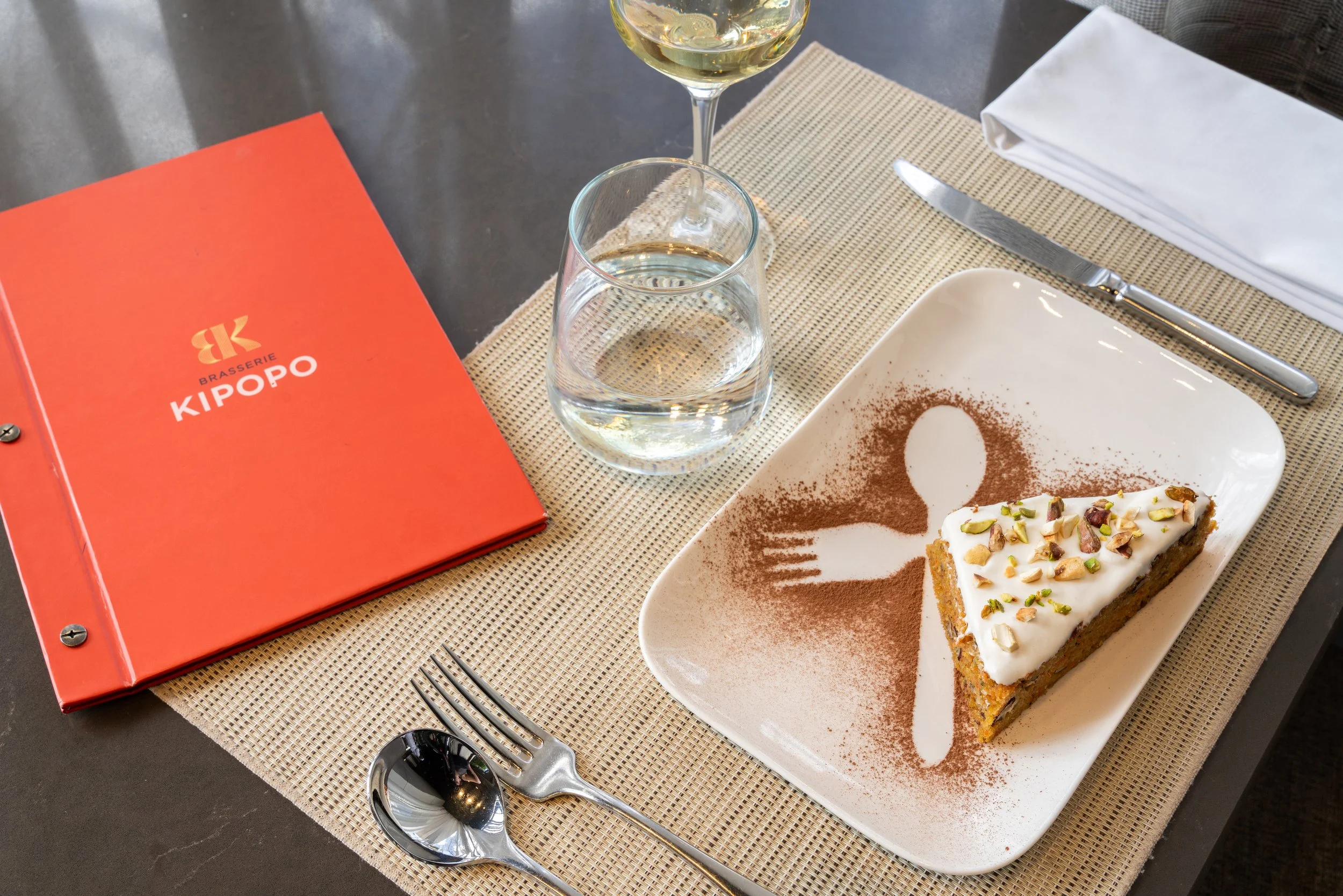 Une table avec un menu rouge de la Brasserie Kipopo, une part de gâteau à la pistache et à la crème, un verre de vin blanc, un verre d'eau, une cuillère, une fourchette et une serviette blanche.