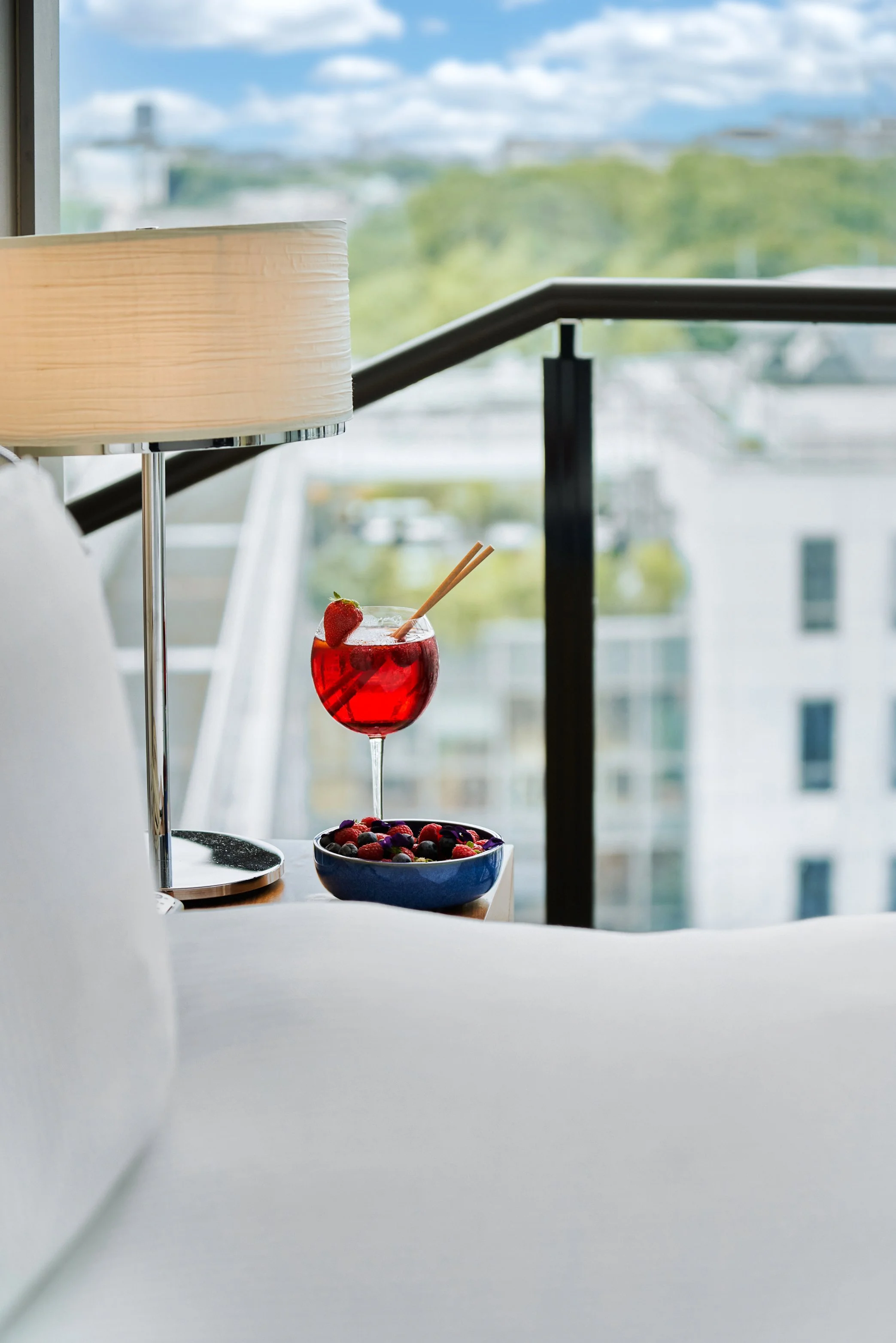 Verre de cocktail rouge garni d'une fraise, assiette de baies mélangées, lampe de chevet et balcon avec vue urbaine en arrière-plan.