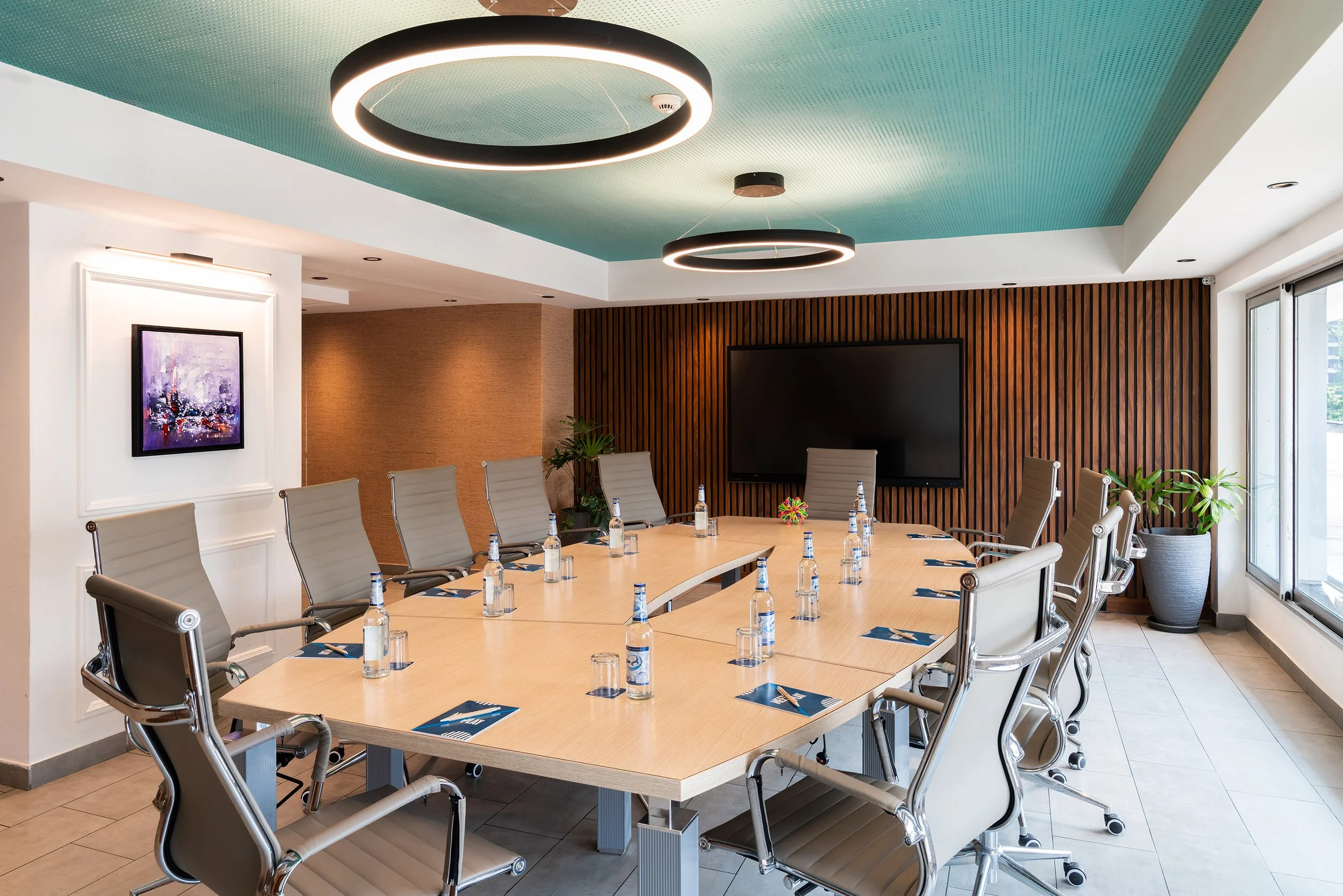 Salle de réunion boardroom avec une grande table, chaises en cuir, bouteilles d'eau, et un grand écran télévision sur un mur en bois