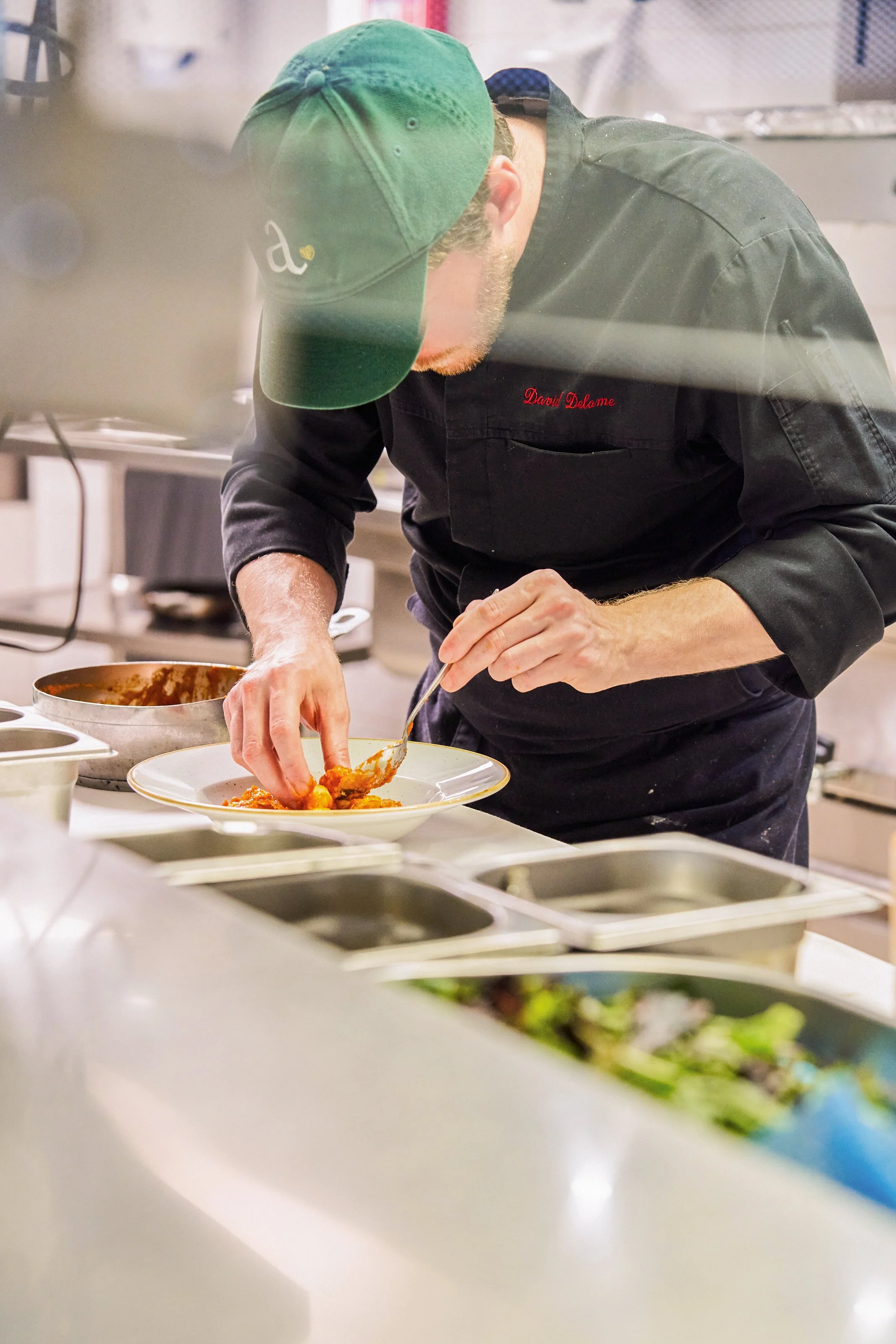 Chef de cuisine, portant une casquette verte, servant un plat de nourriture dans une cuisine professionnelle.