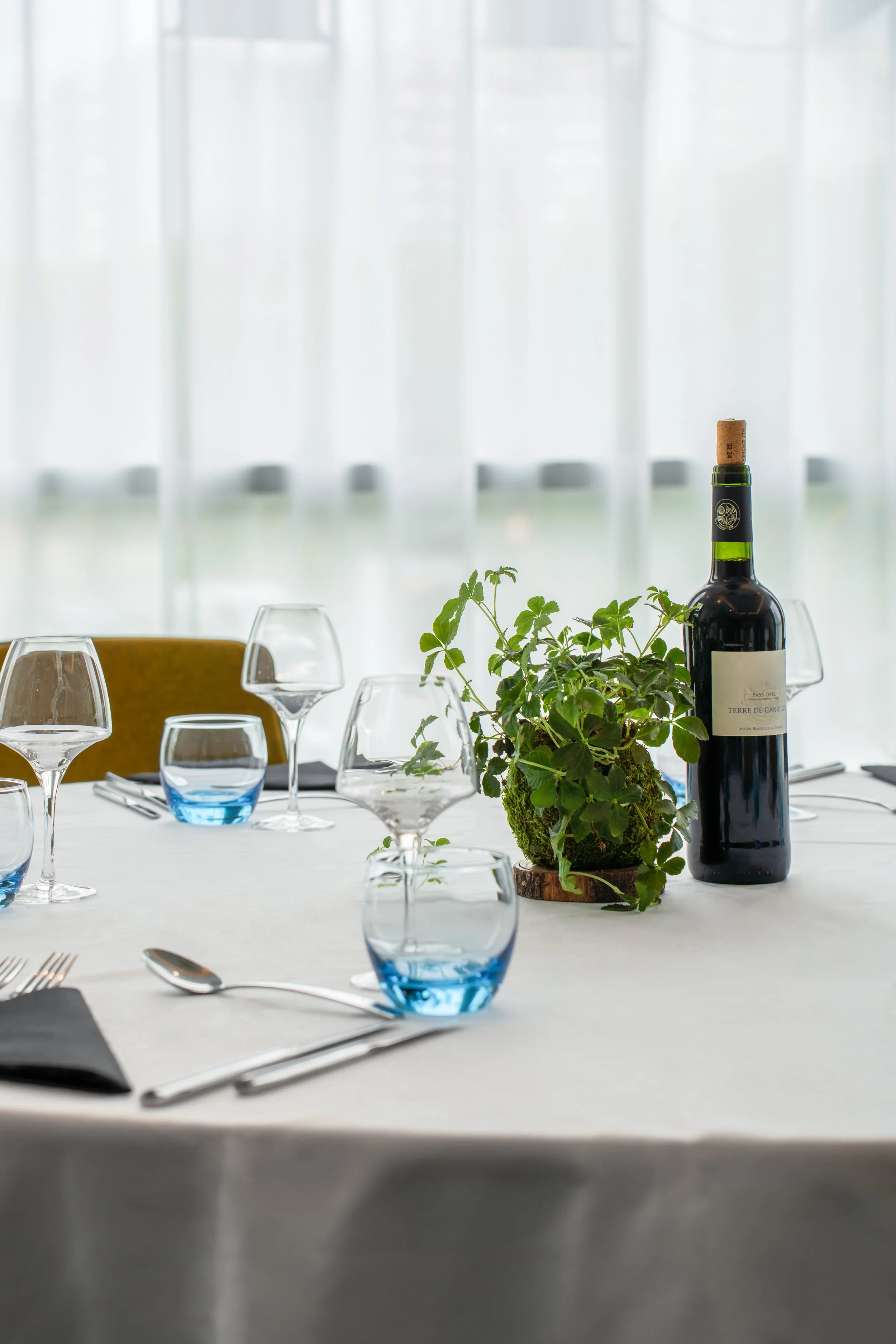 Table de restaurant élégante avec verres à vin et à eau, une bouteille de vin et un bouquet de plantes vertes, lumière naturelle à l'arrière-plan.