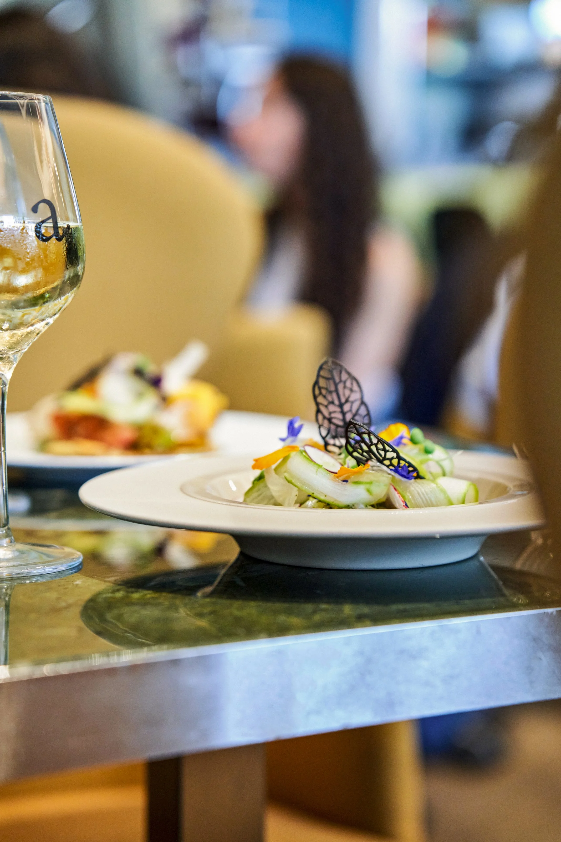 Assiette de salade avec des feuilles vertes, des fleurs comestibles et des décorations en forme de papillons, une coupe de vin blanc à côté, dans un restaurant élégant.