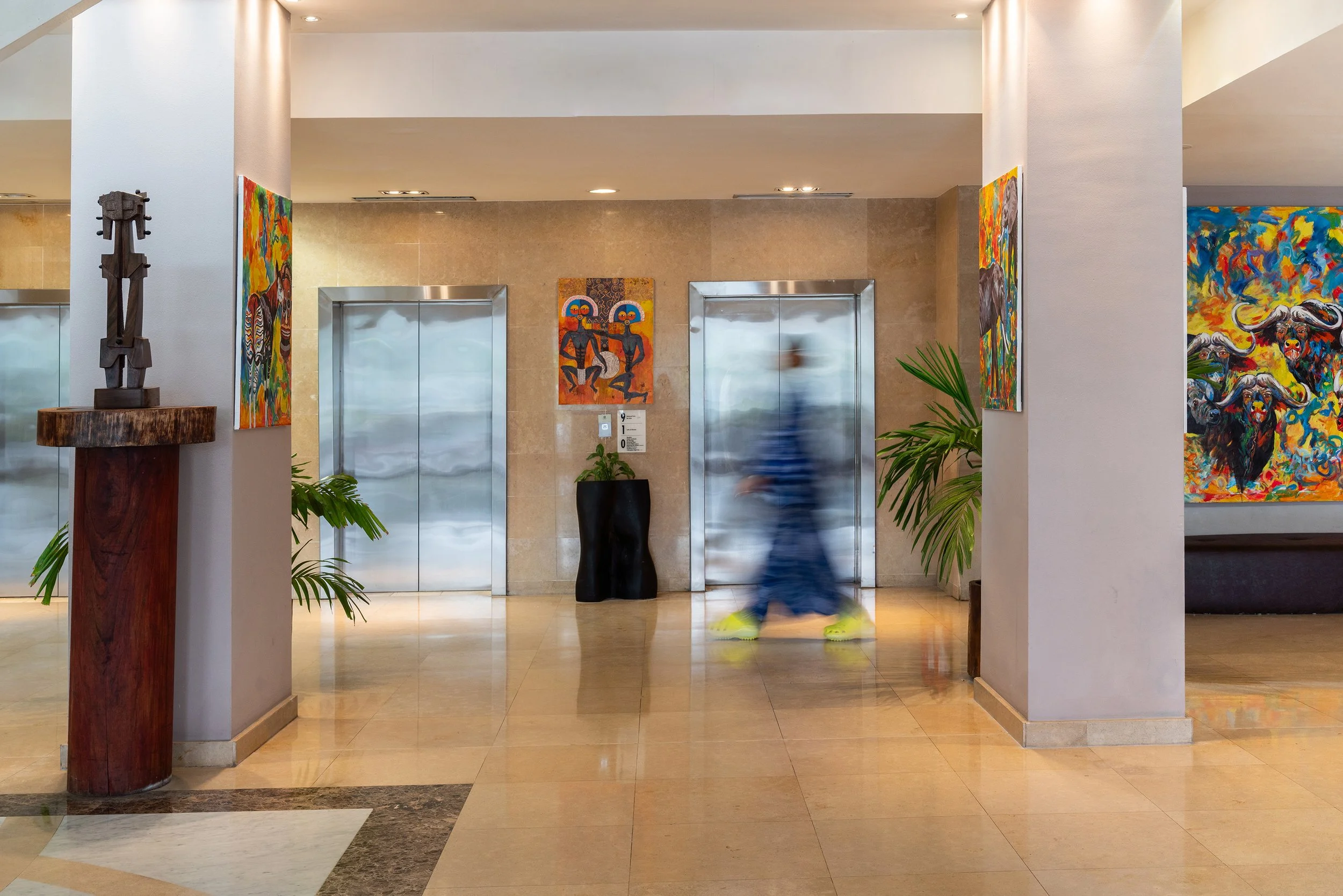 Halle d'ascenseur dans un hall d'hôtel avec des peintures colorées sur les murs et des plantes vertes, une femme en mouvement passant devant les ascenseurs.