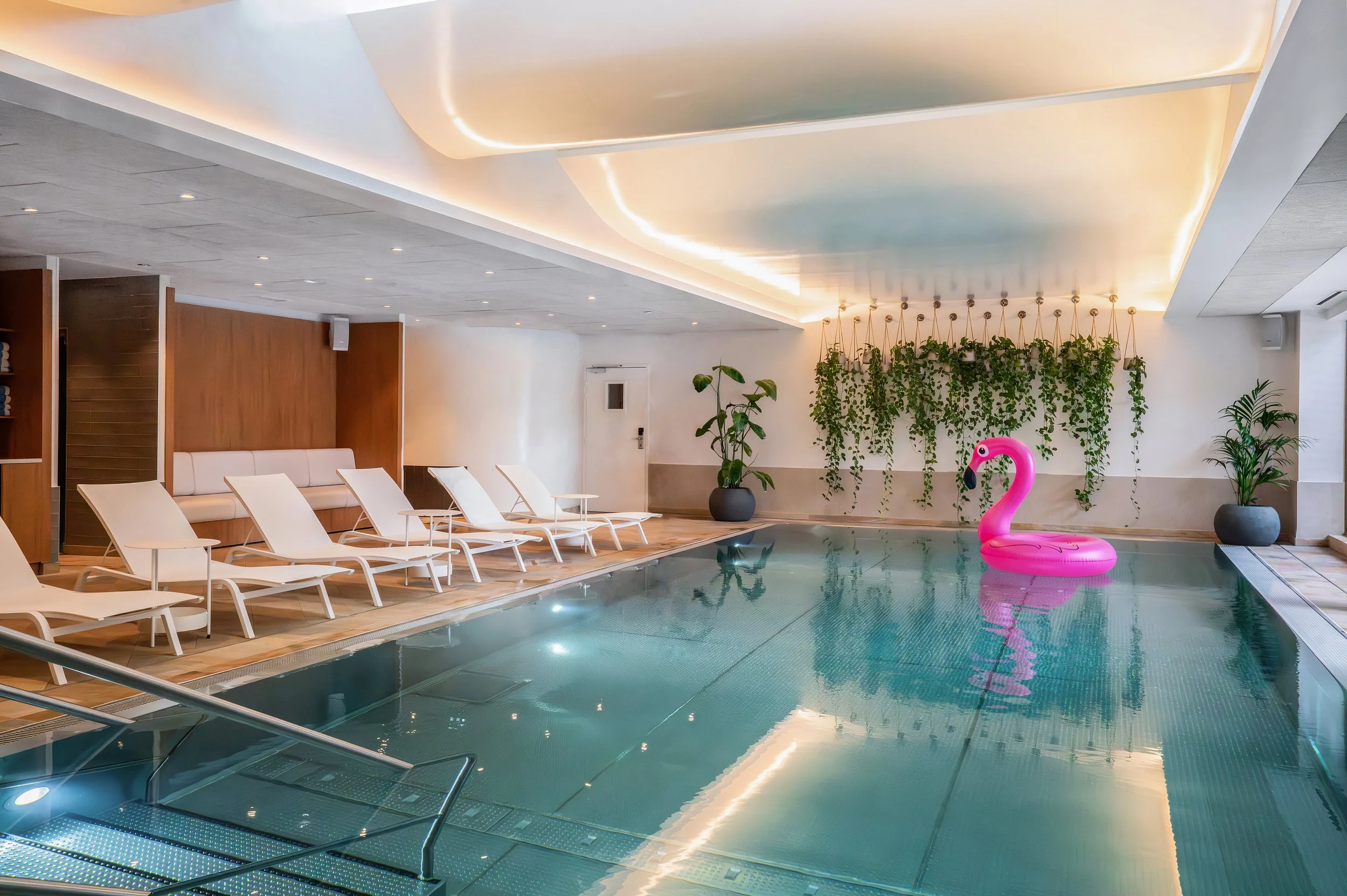 Piscine intérieure avec une bouée en forme de cygne rose, sièges loungers blancs, plantes vertes, décoration moderne, éclairage tamisé.