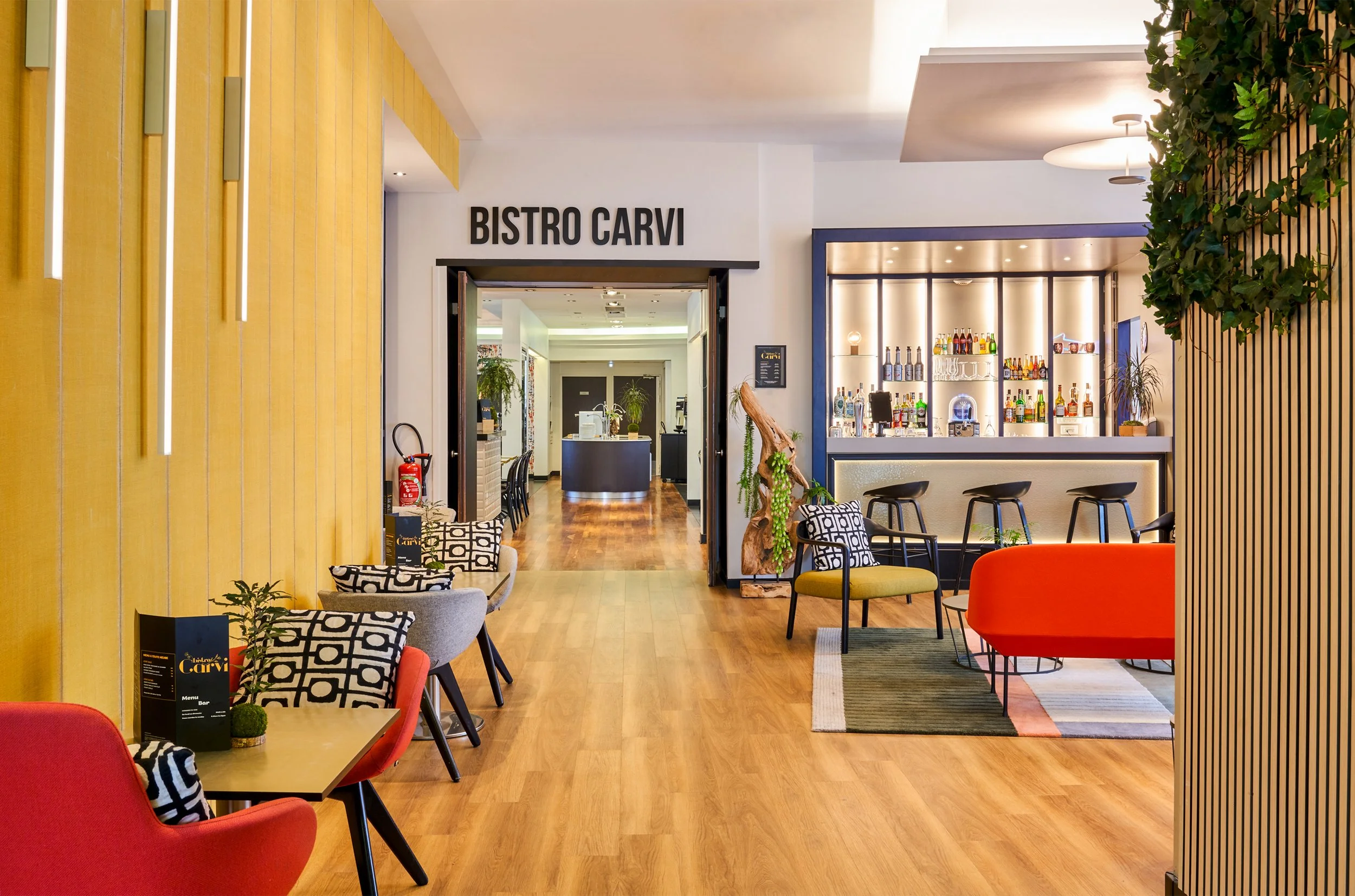 Intérieur d'un bar avec des meubles modernes, des fauteuils colorés, un bar avec des bouteilles et une décoration végétale.