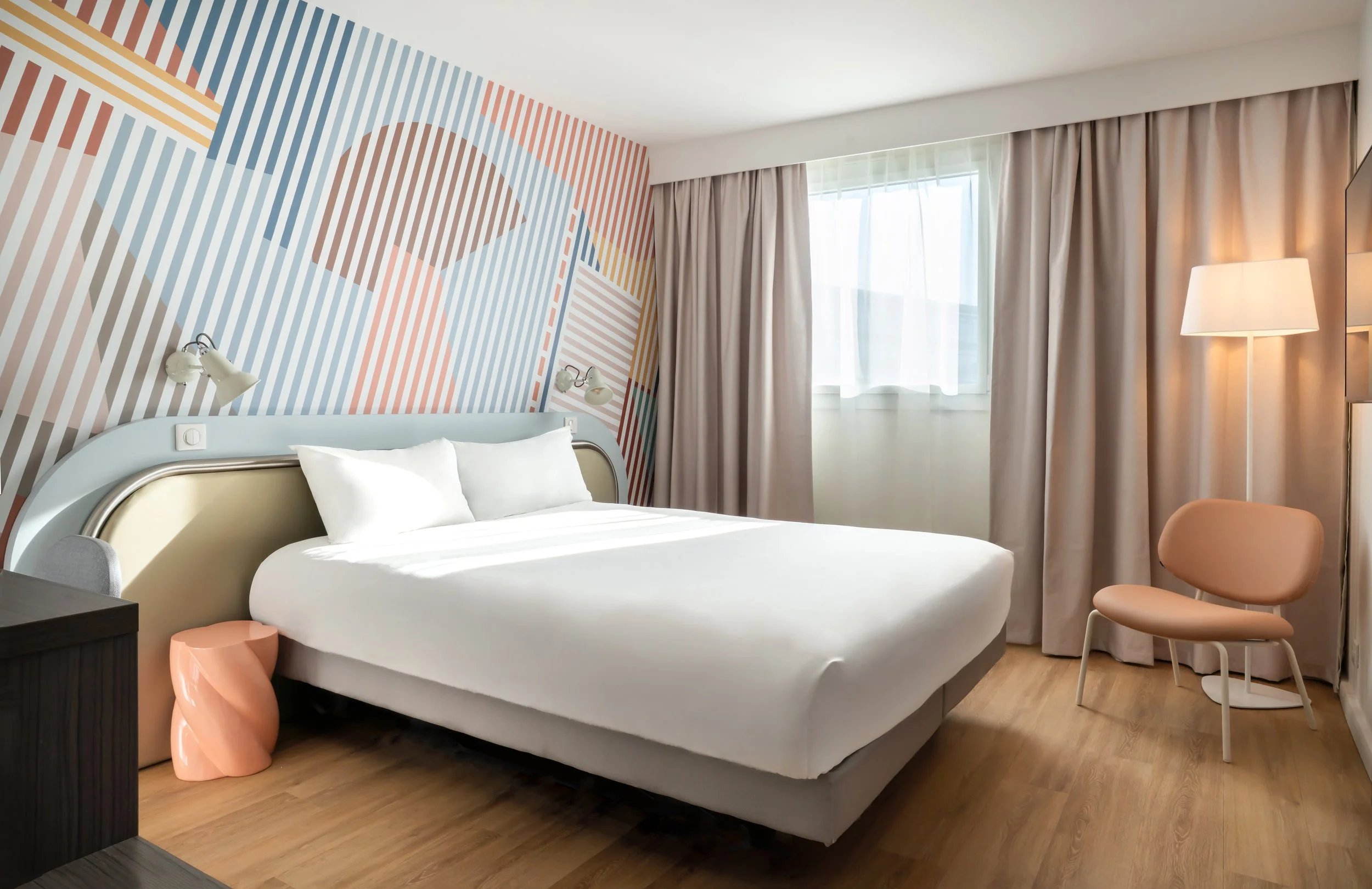 Chambre d'hôtel avec mur d'accent coloré, lit blanc, lampes murales, chaise beige, lampadaire et fenêtres avec rideaux beige.