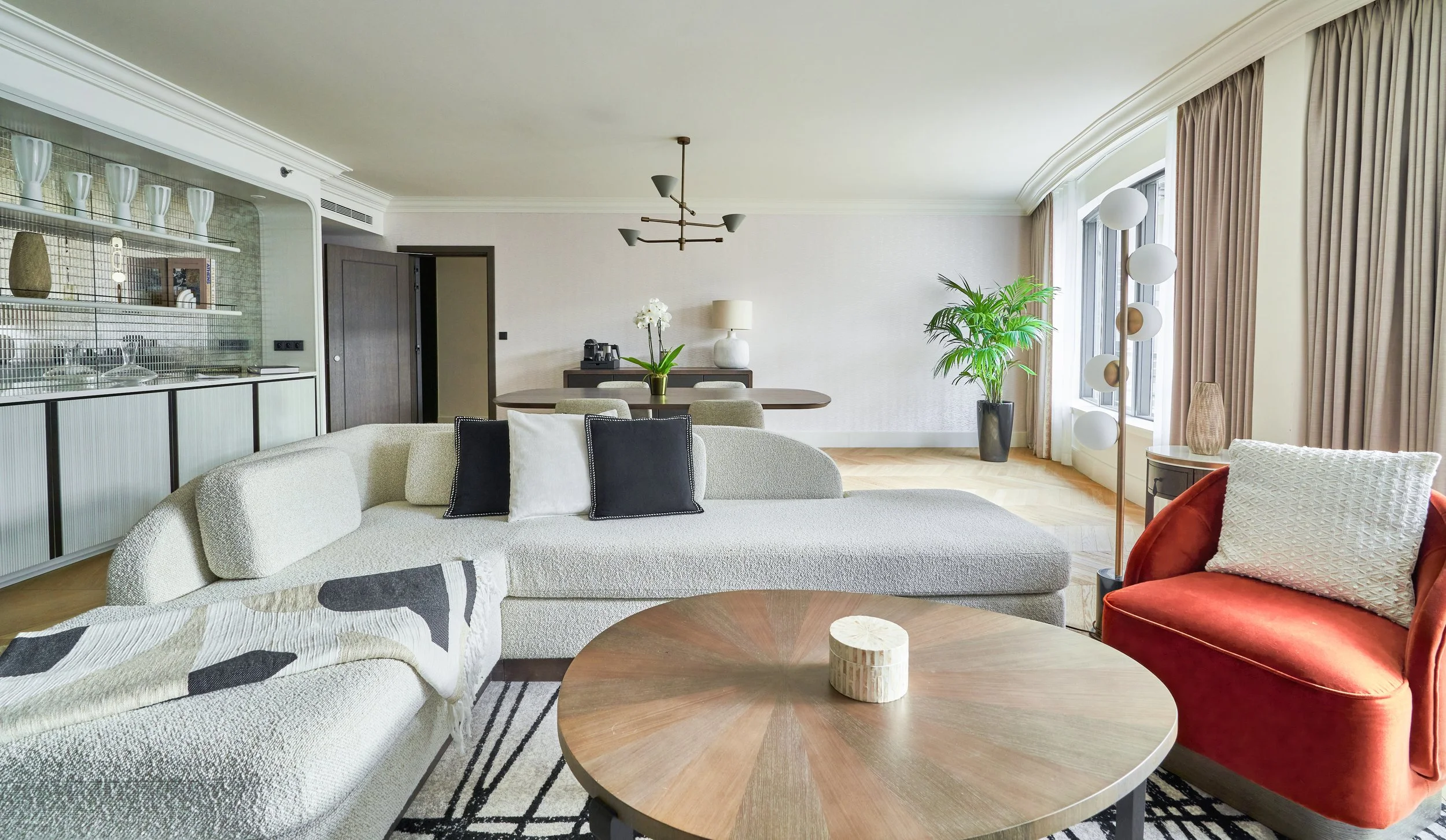 Salon spacieux d'une suite avec canapé beige, fauteuil rouge, table en bois, lampadaire moderne, grande fenêtre avec rideaux beiges, plantes vertes, décor élégant et moderne.