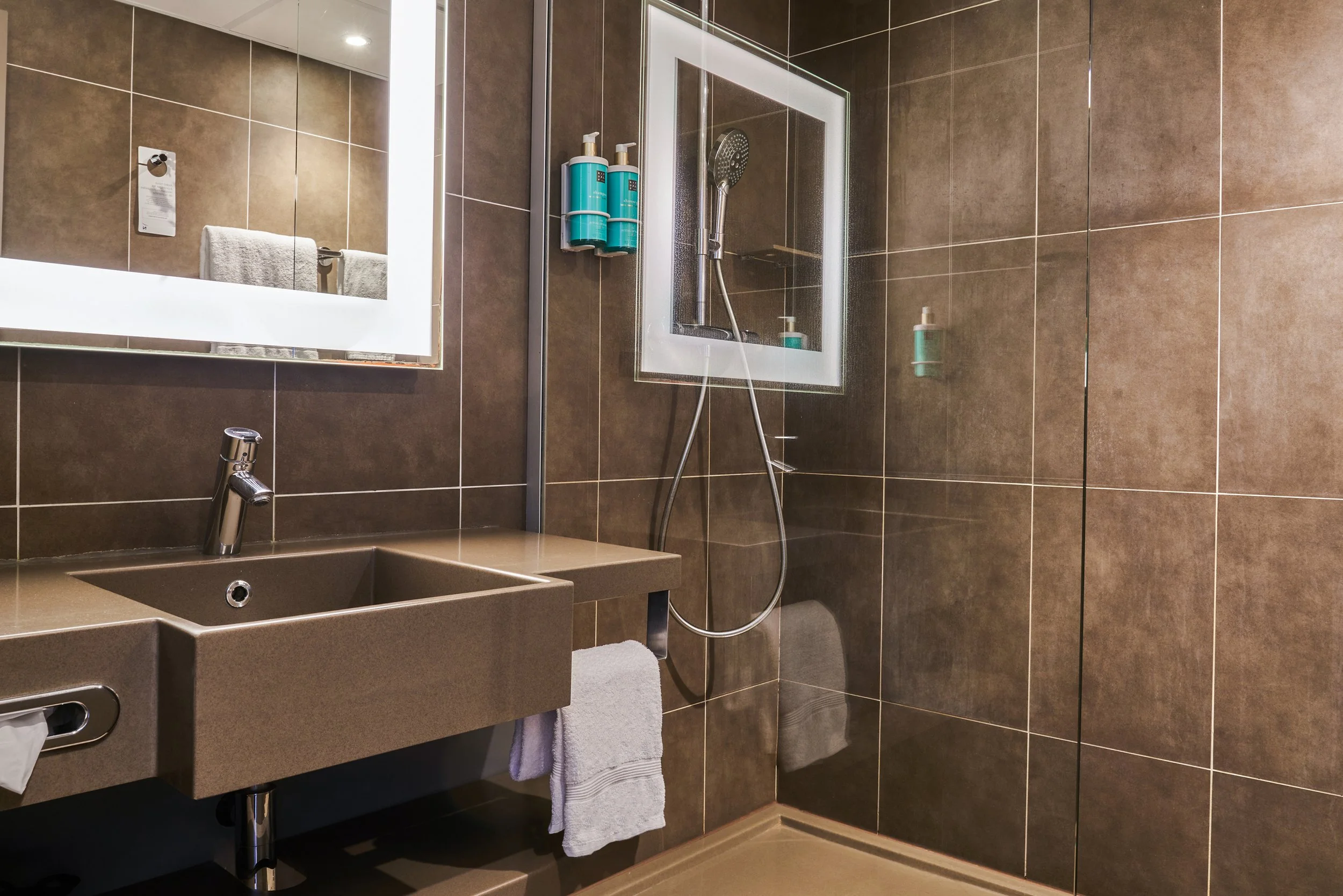 Salle de bain avec mur et lavabo en carrelage marron, miroir avec éclairage LED, douche avec paroi en verre, distributeurs de savon et produits de toilette, serviette blanche à côté du lavabo.