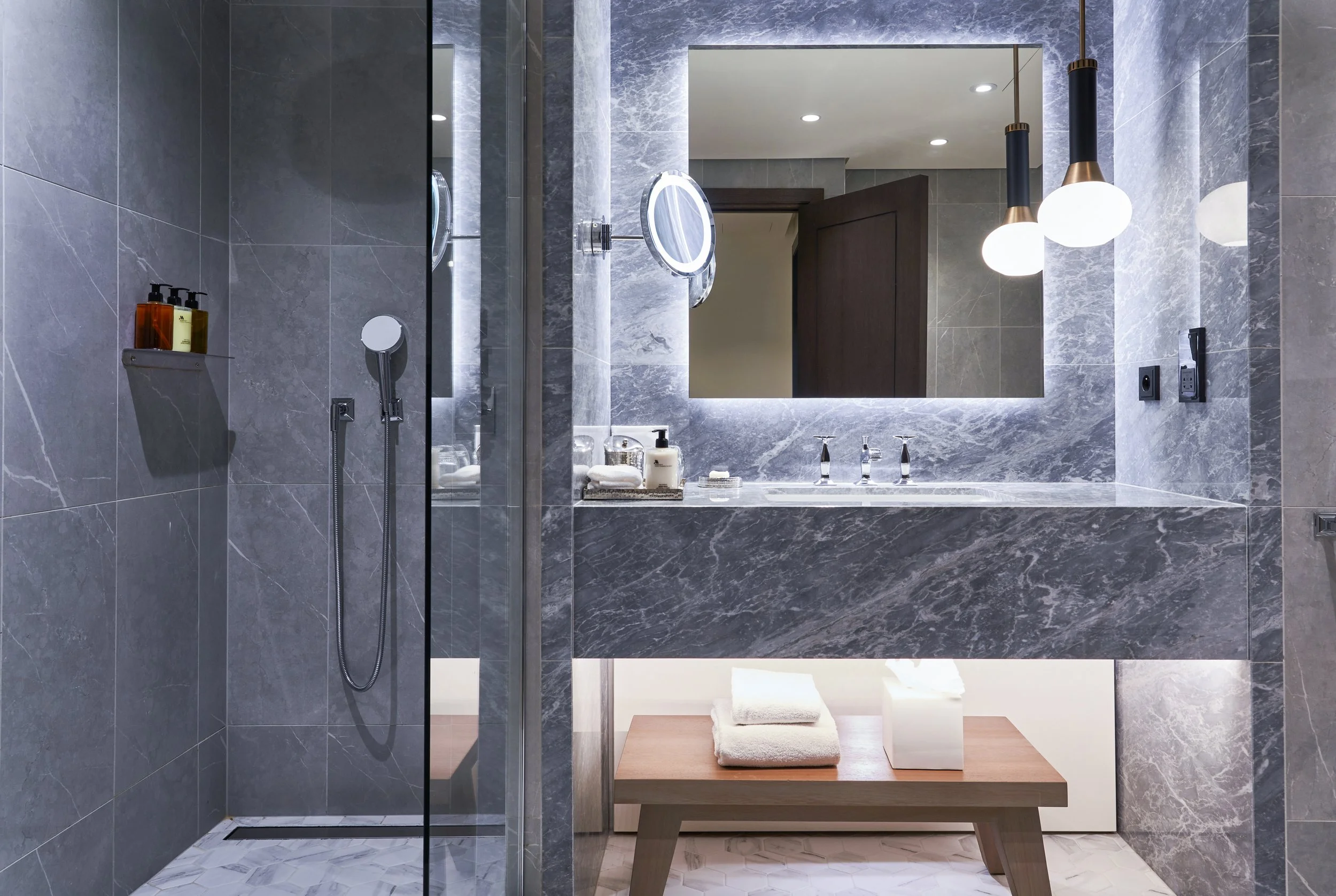 Salle de bain moderne avec paroi vitrée, miroir éclairé, lavabo en marbre, éclairage suspendu, et accessoires de toilette.