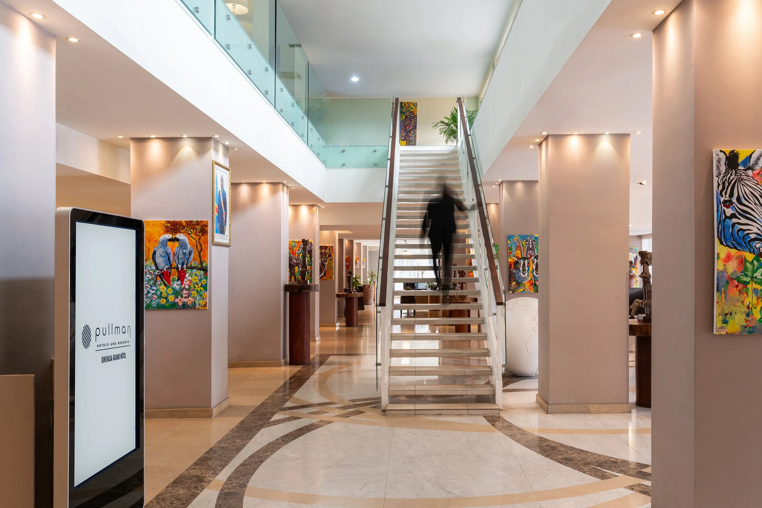 Intérieur d'un hôtel moderne avec une réception, des œuvres d'art colorées au mur, un escalier central, et un sol en marbre.