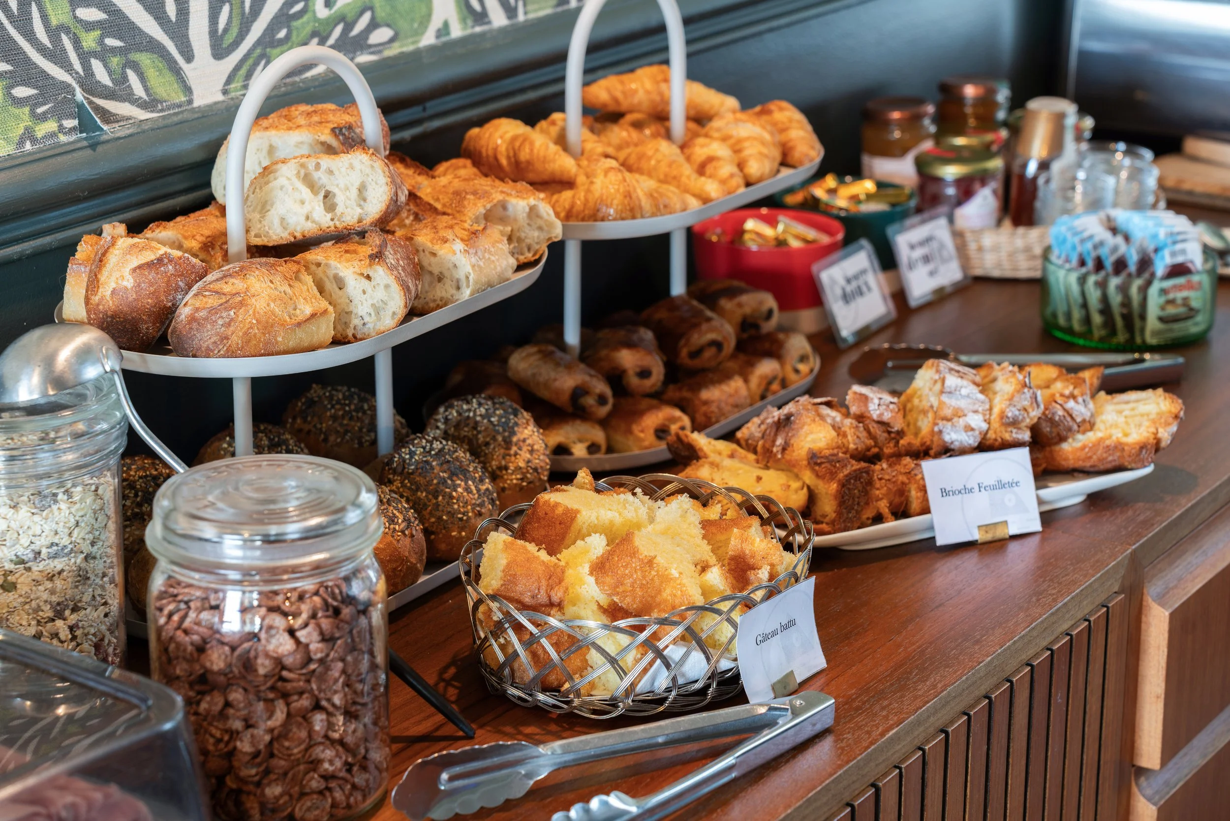Sélection de viennoiseries françaises sur un buffet, comprenant des croissants, des pains au chocolat, des tartes, et des gâteaux, avec des pots de céréales et autres condiments en arrière-plan.