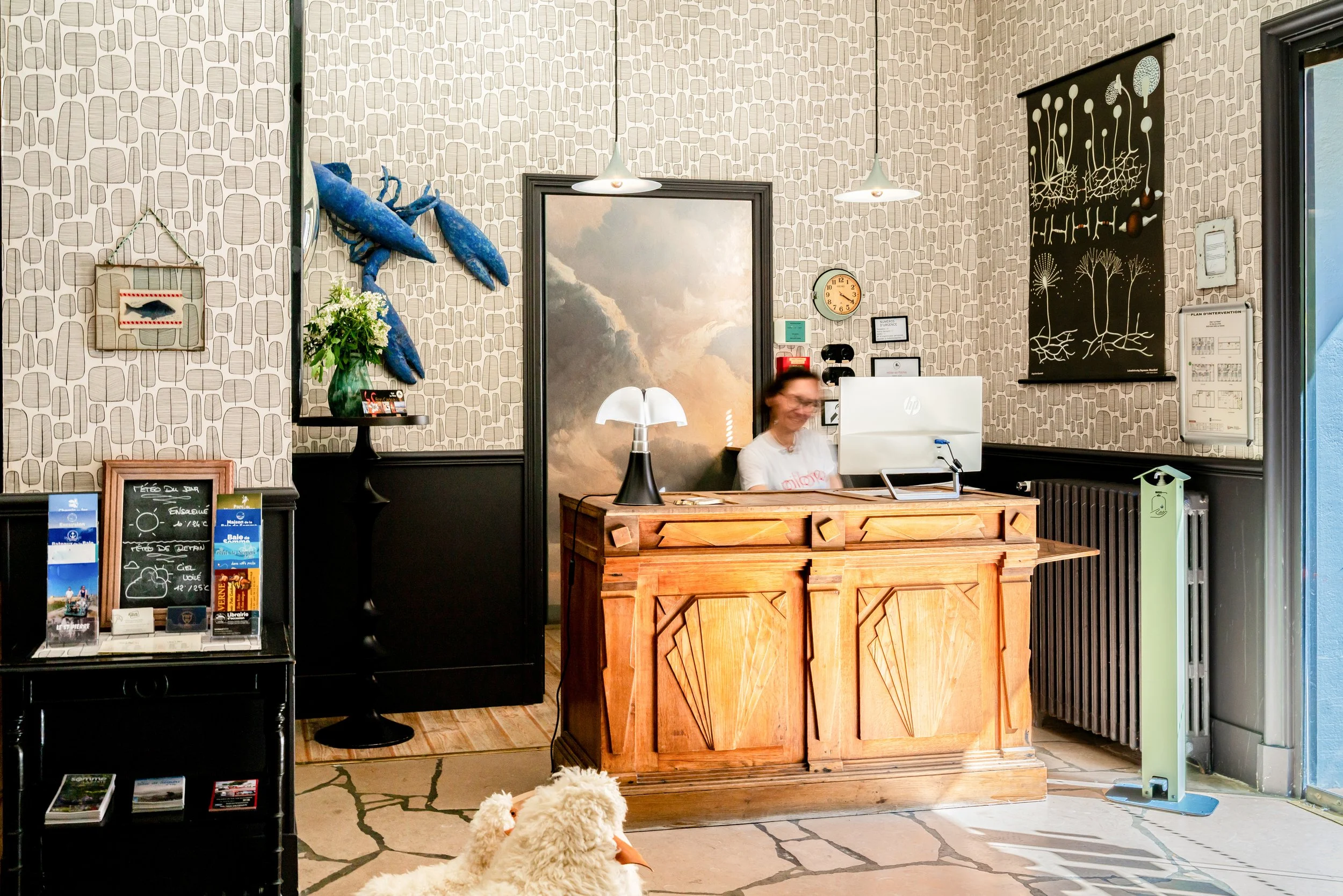 Récéption d'hôtel avec une employée derrière un comptoir en bois, un ordinateur et une lampe. Mur avec décorations de poissons en laine, dessins de champignons et silhouettes d'arbres. Canard blanc en peluche au premier plan et petits panneaux d'info