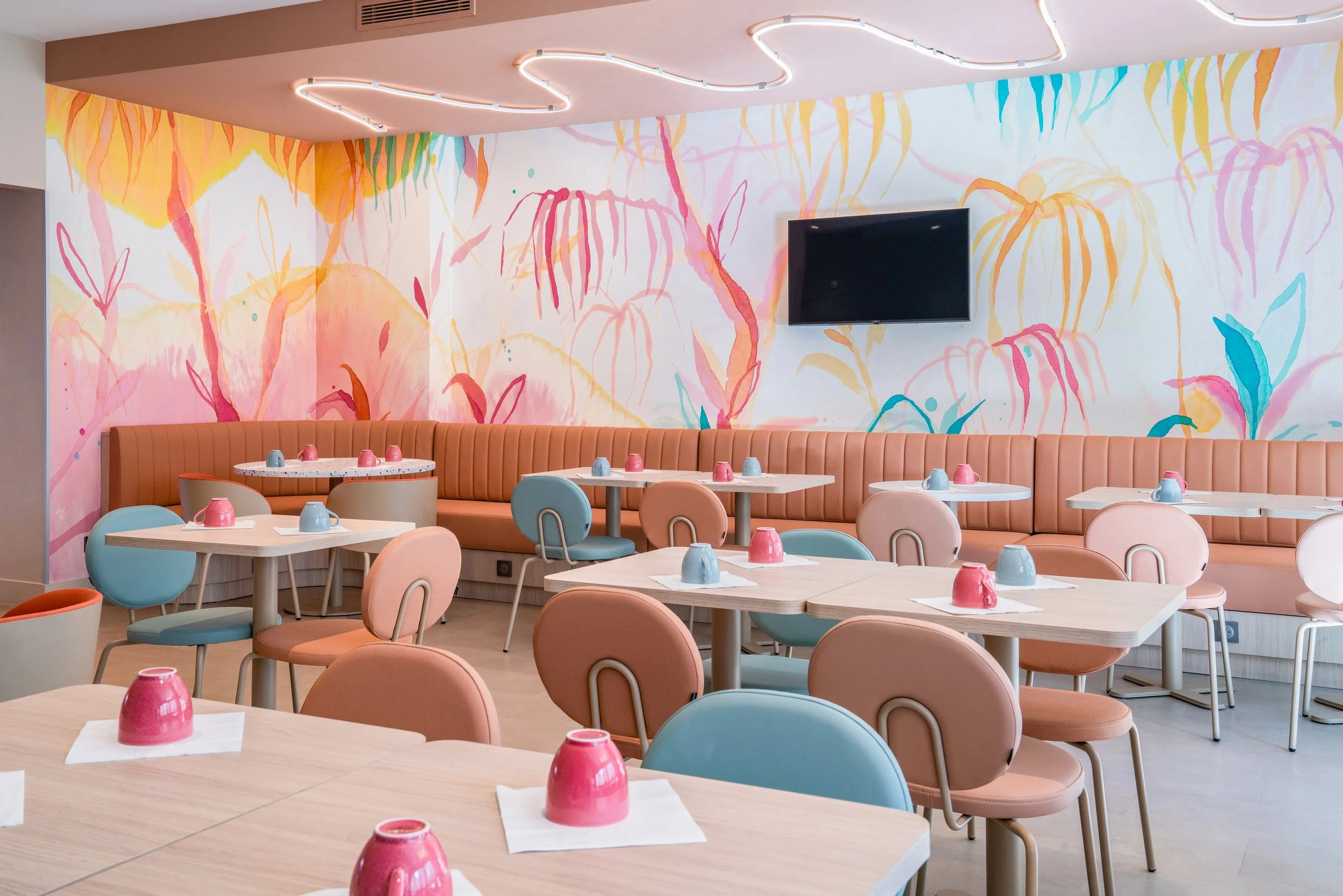Salle à manger au décor pastel avec chaises roses, bleues et beiges, tables avec assiettes colorées, mural avec peinture florale abstraite, télévision allumée au mur.