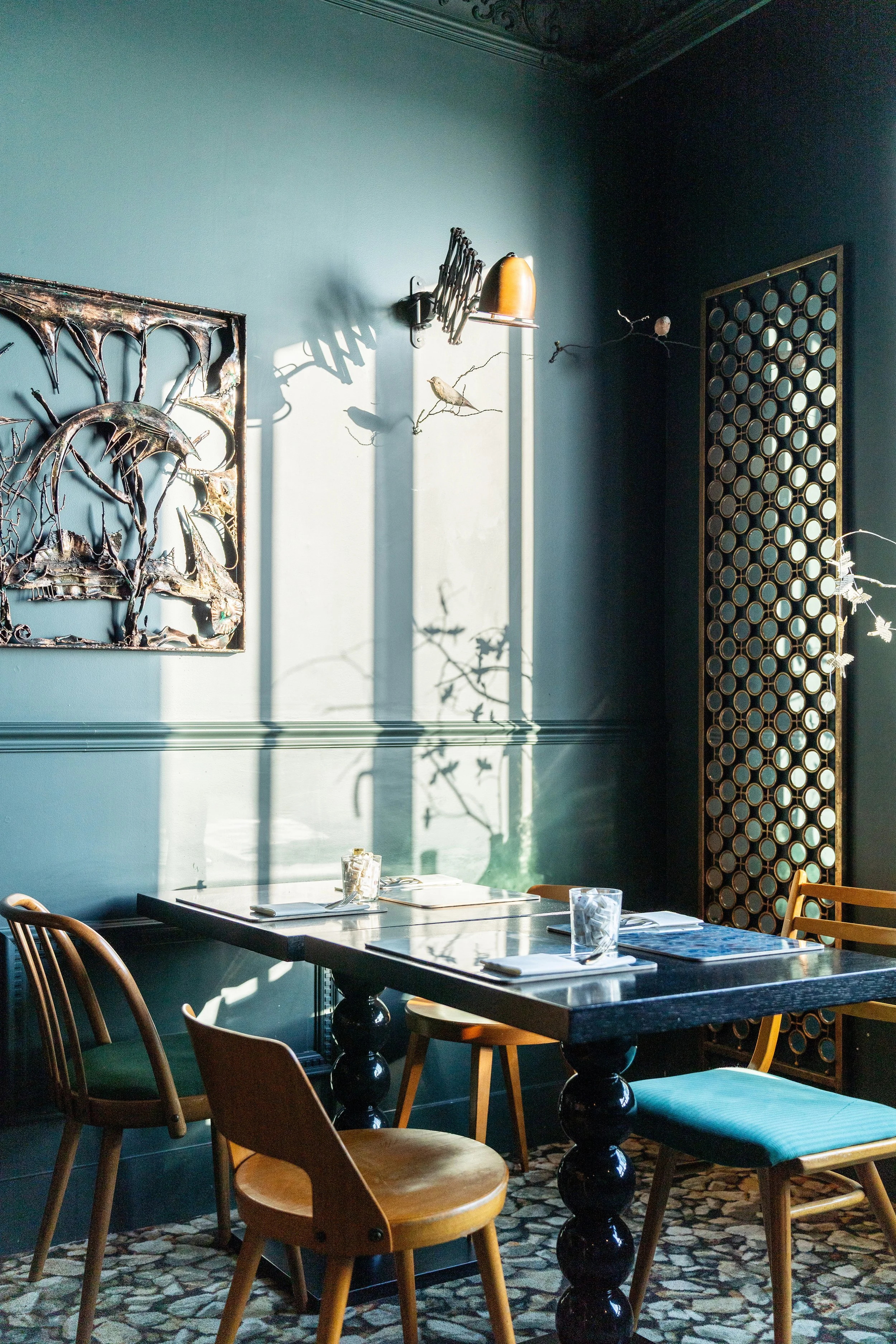 Intérieur d'une salle à manger avec une table en marbre noir, quatre chaises en bois de styles différents, et une décoration murale artistique, avec du soleil qui projette des ombres sur le mur bleu.