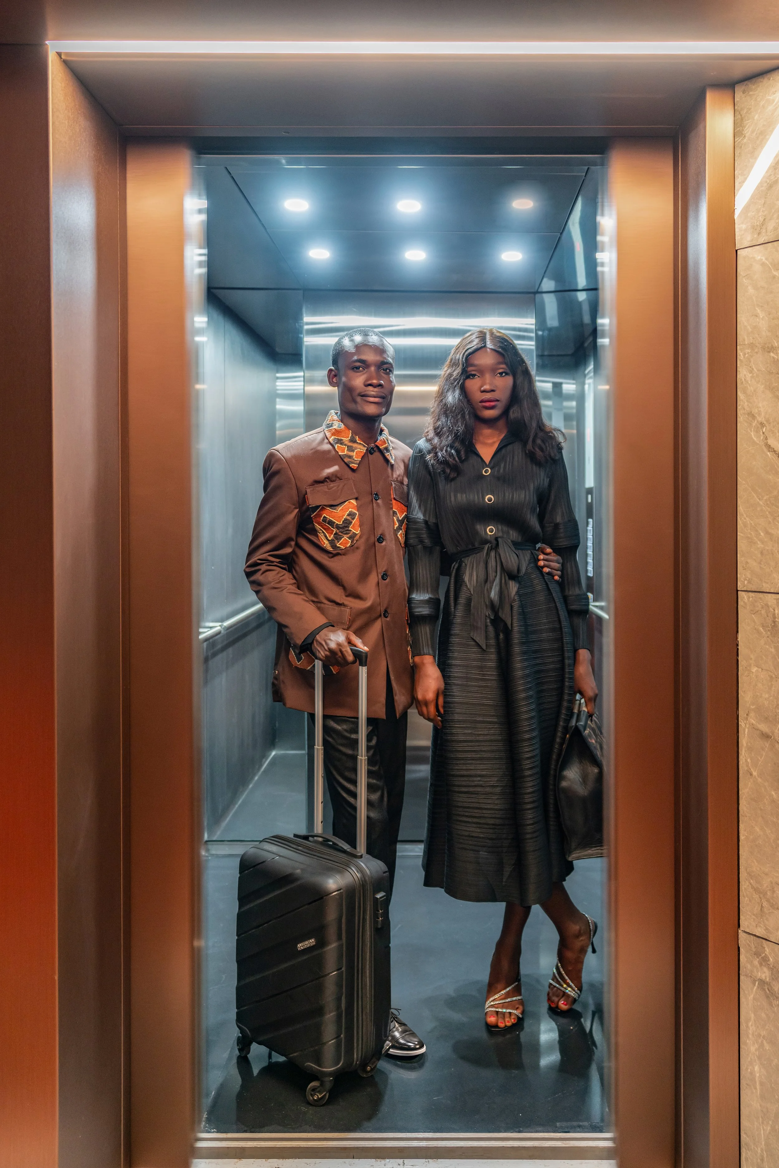 Un couple élégant se tient dans un ascenseur, l'homme avec une valise et la femme avec un sac, tous deux habillés de façon chic, dans un hôtel moderne.
