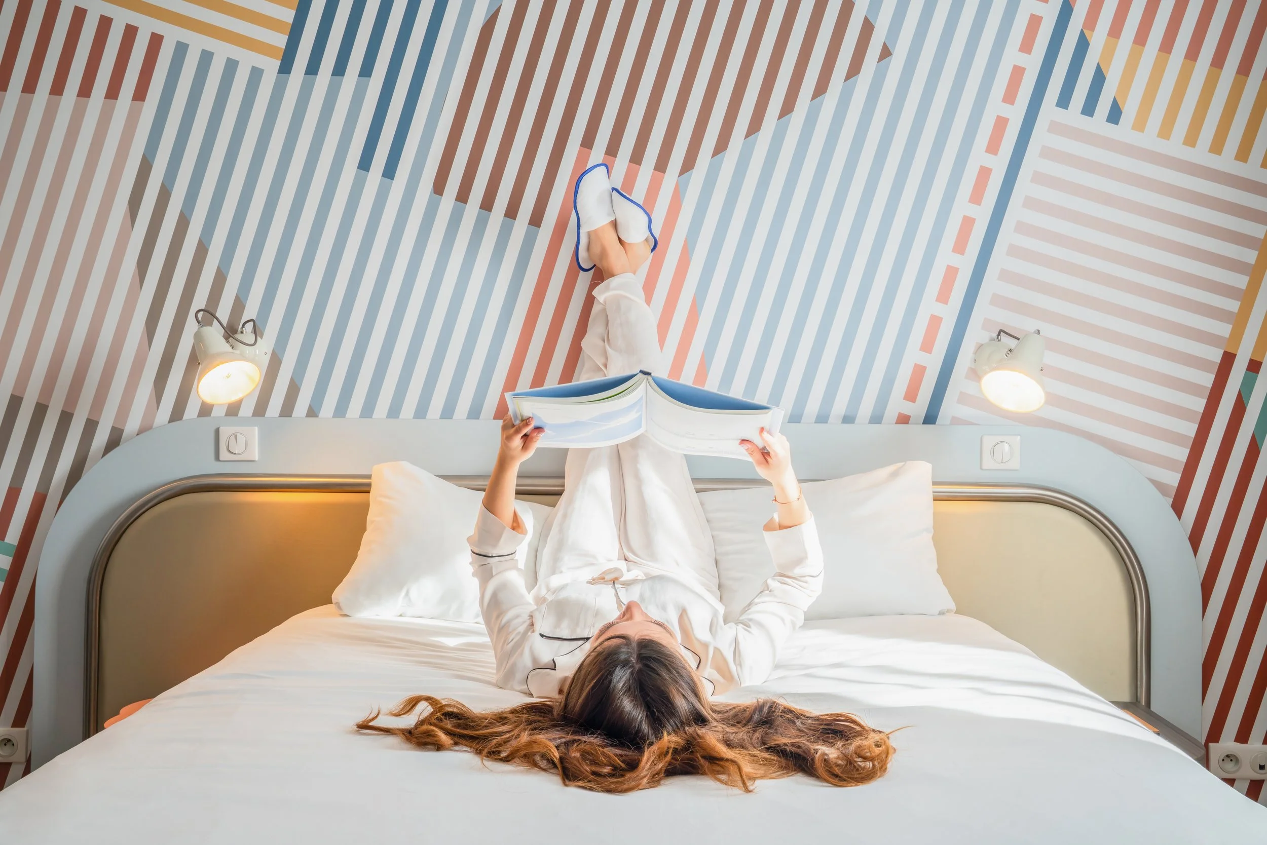 Une femme en pyjama blanc lit un magazine sur un lit blanc, couchée dos, dans une chambre avec un mur coloré à motifs géométriques et deux lampes de chaque côté du lit.